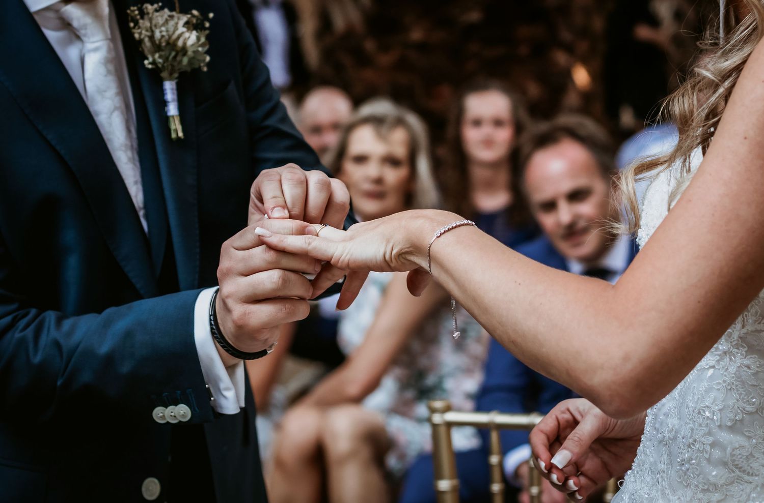 Close-up of bride placing a wedding ring on grooms finger with guests watching in the background.