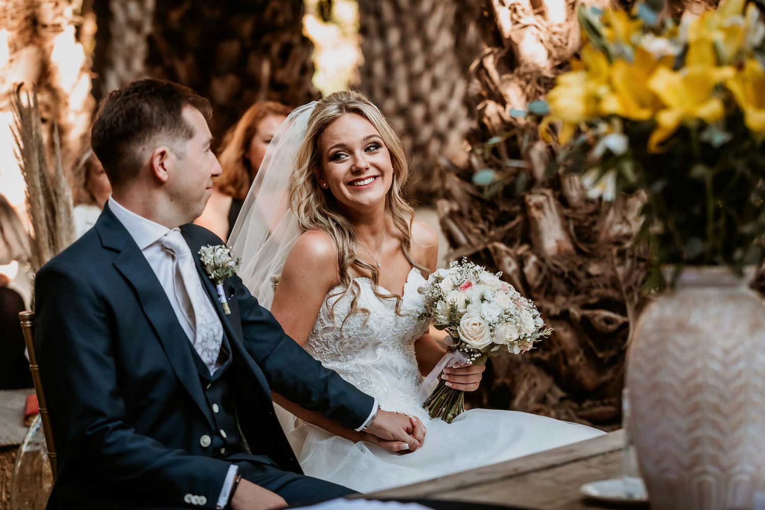 Bride and groom sitting together at outdoor wedding ceremony altar with floral bouquet.