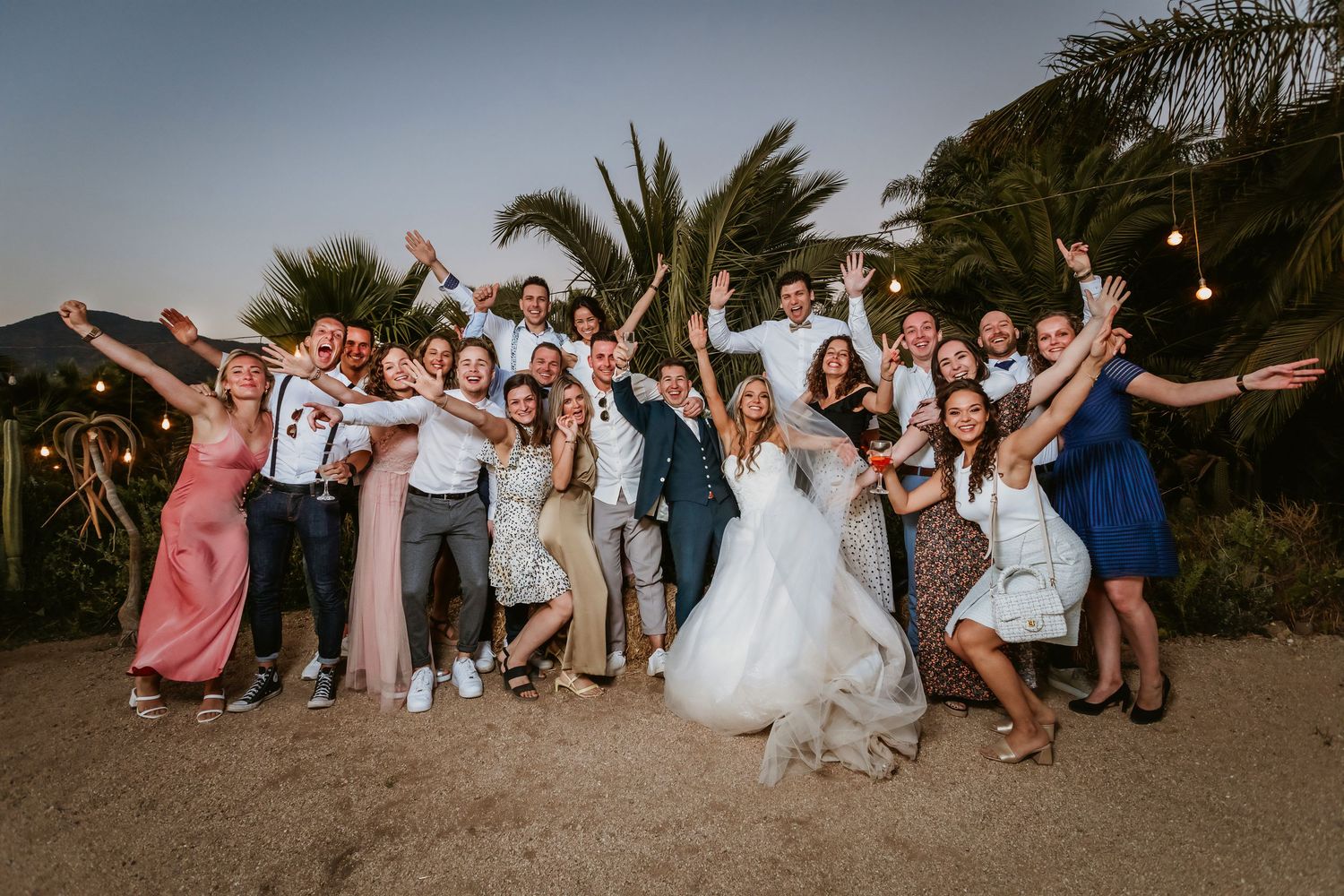 Large joyful wedding party group photo celebrating outdoors on sandy ground with palm trees.