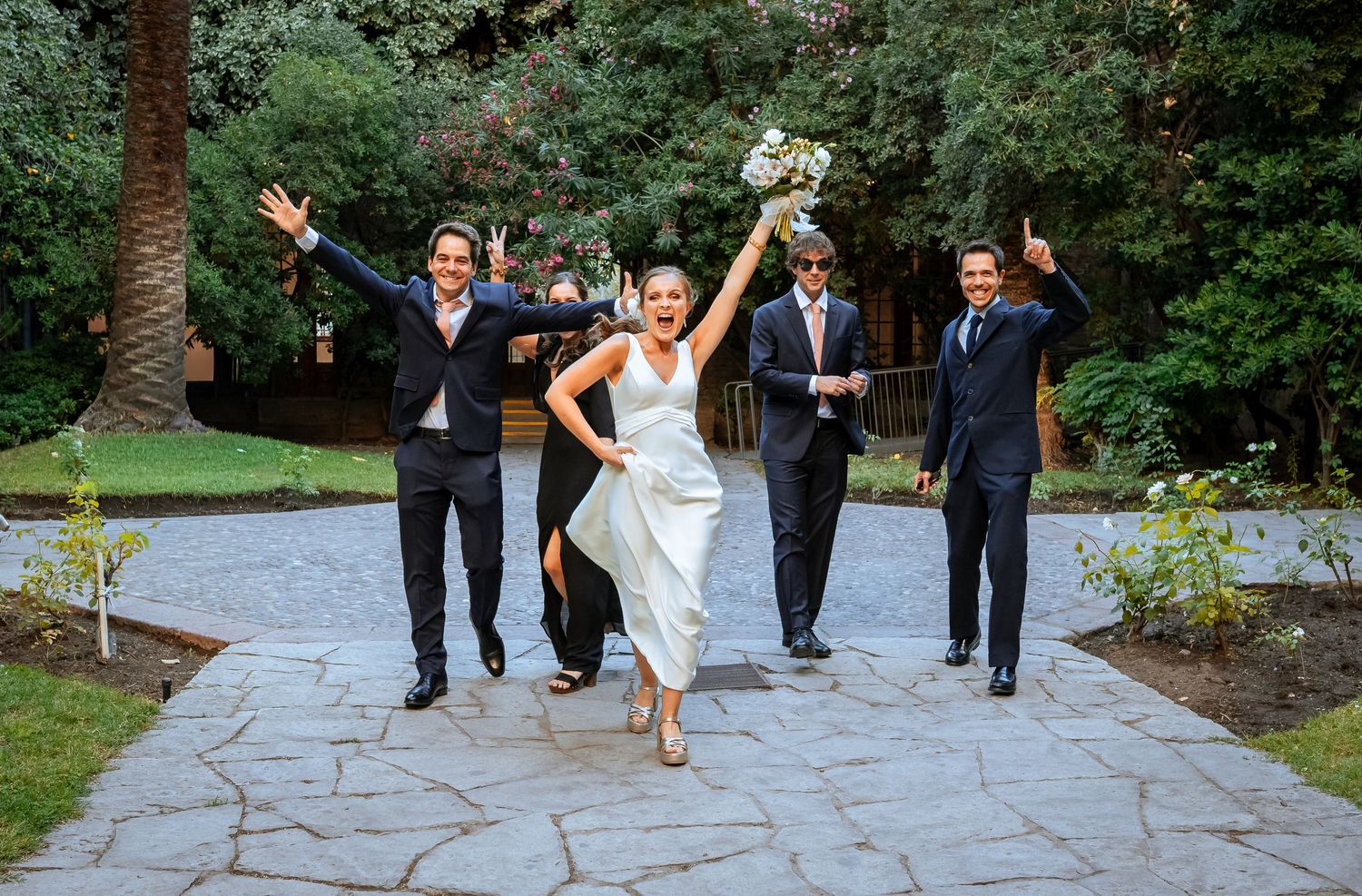 Bride in white dress running joyfully with wedding party on stone pathway outdoors.