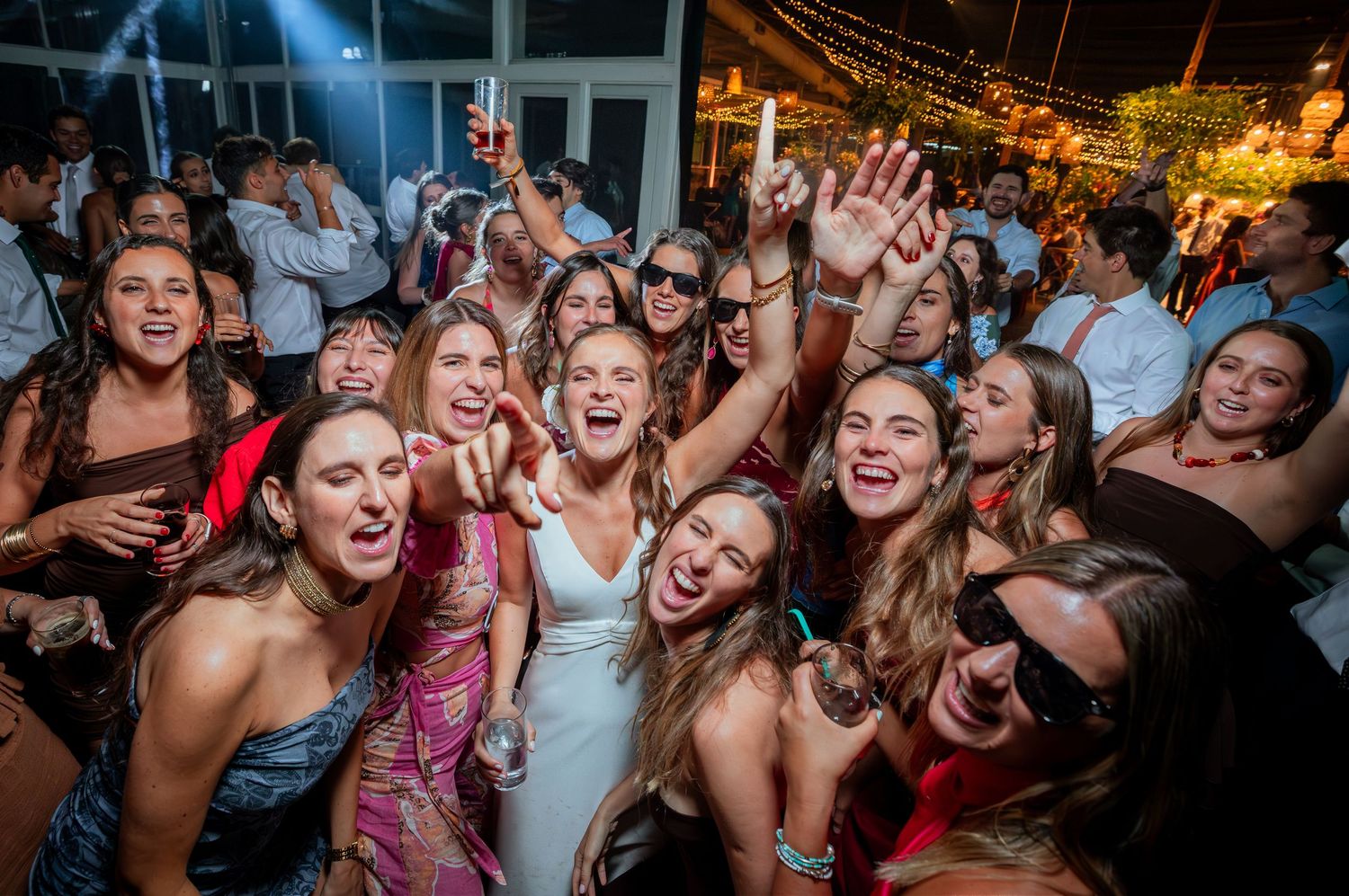 Smiling bride in white dress poses with large group of joyful wedding guests at outdoor reception.