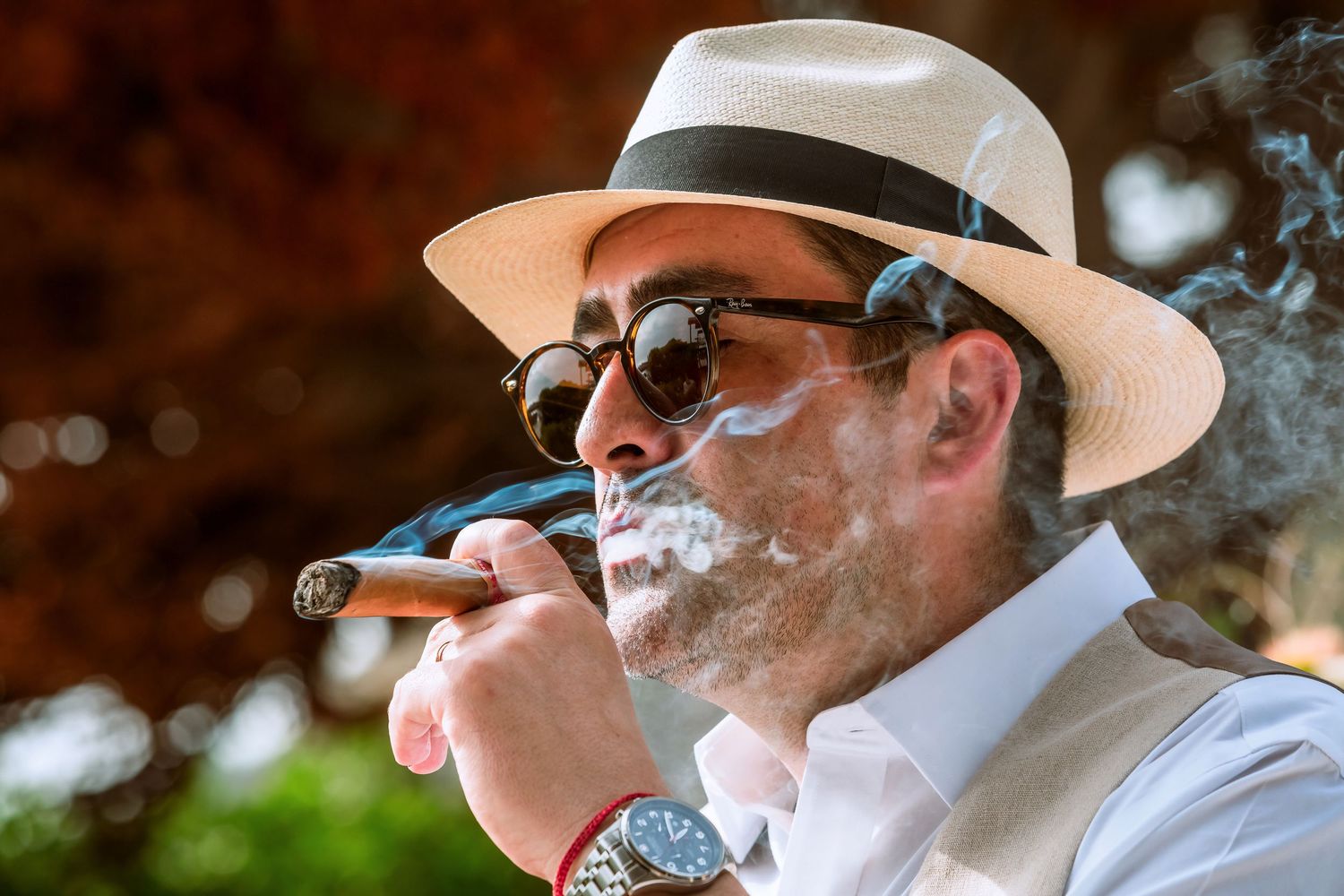 Close-up of man in panama hat exhaling cigar smoke, wearing sunglasses and a luxury watch outdoors.