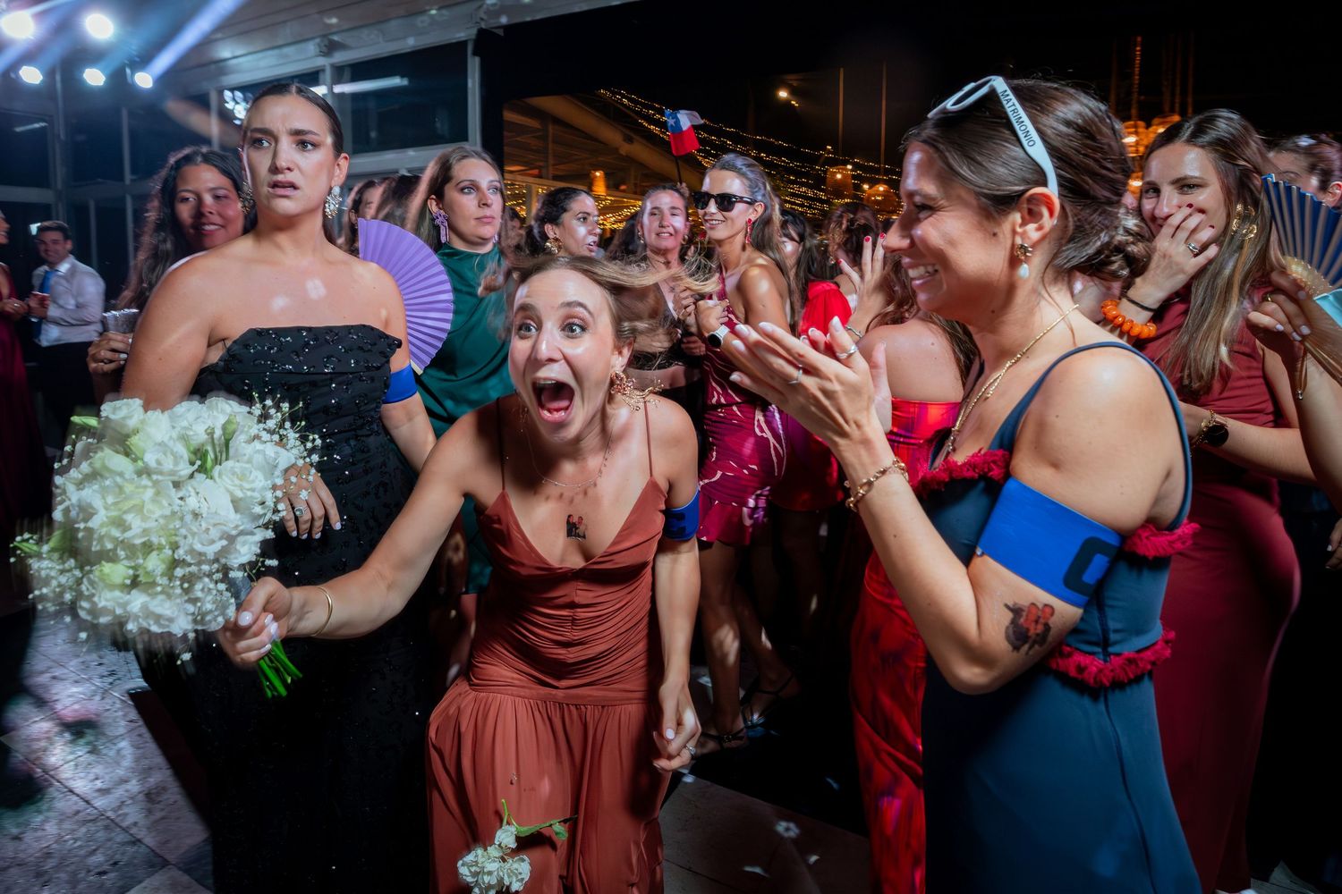 Excited wedding guests react with joy as the bride tosses her white bouquet at a lively reception.