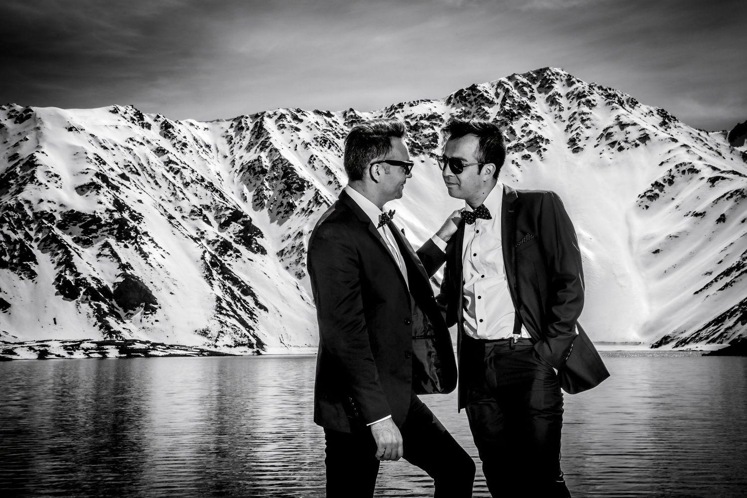 Black and white close-up of two grooms in tuxedos sharing an intimate moment by a mountain lake.