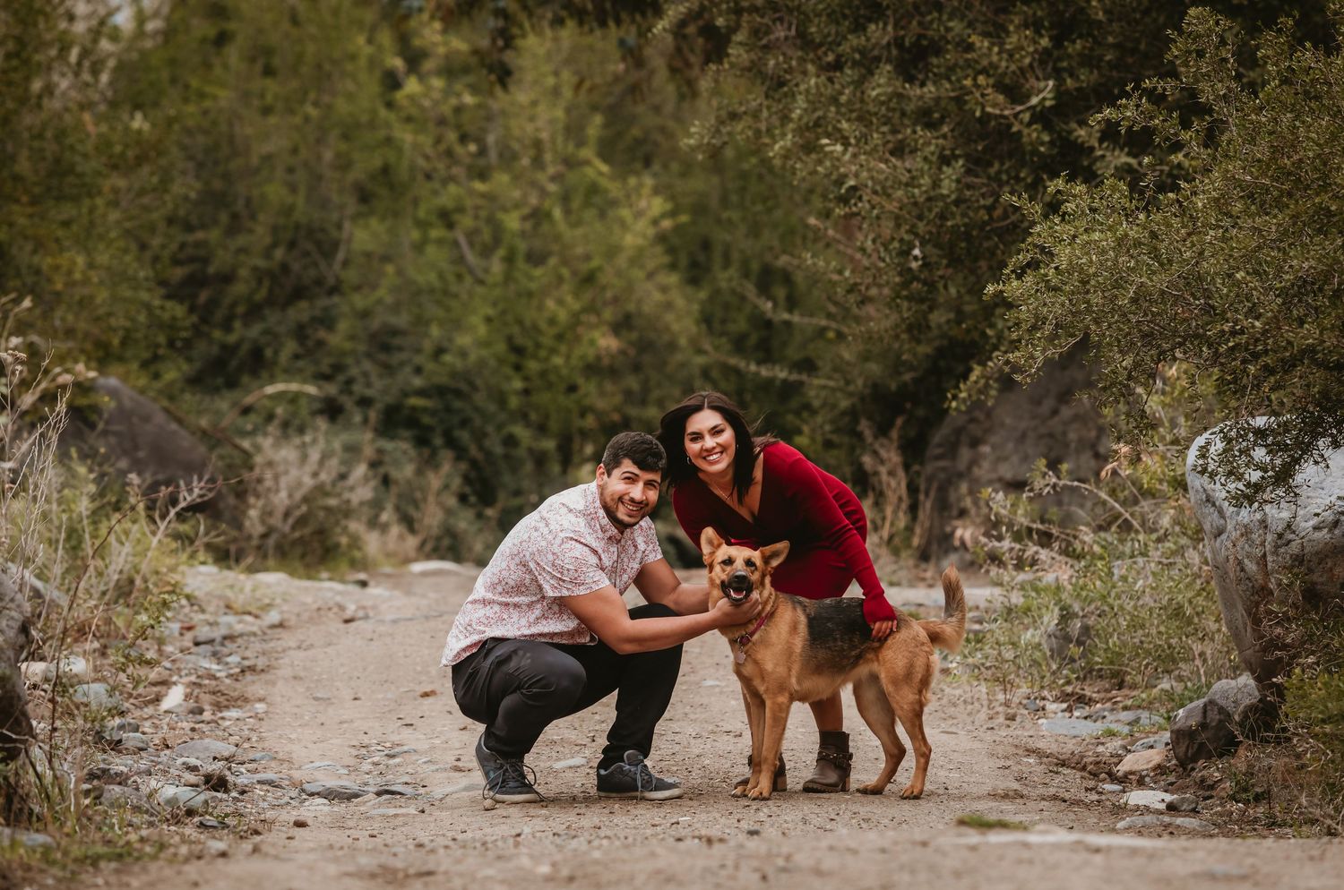 Smiling couple crouching together petting their brown dog on a dirt trail outdoors.