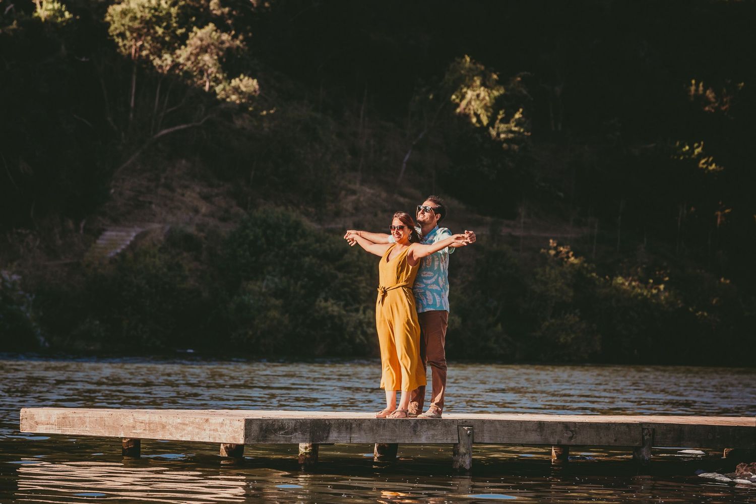 Couple standing on a wooden dock by a lake, woman in yellow dress with arms outstretched.