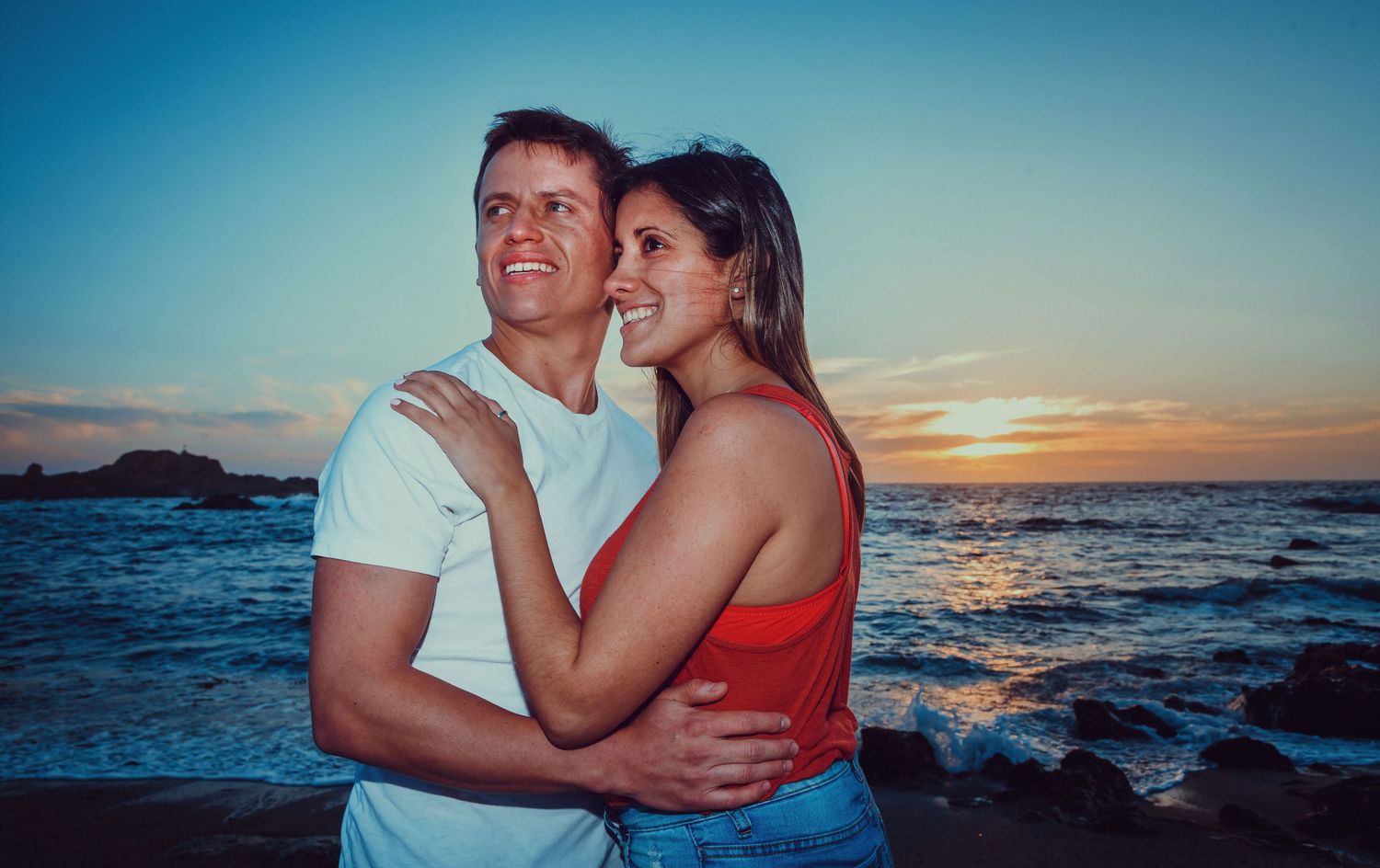 Happy couple embracing and smiling on a rocky beach during a beautiful blue and orange sunset.