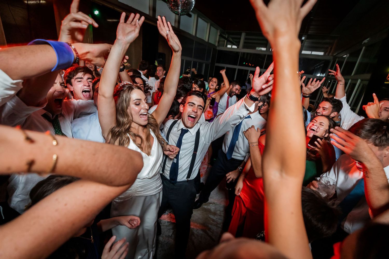 Happy bride and groom celebrating with raised arms surrounded by cheering guests at lively wedding party.
