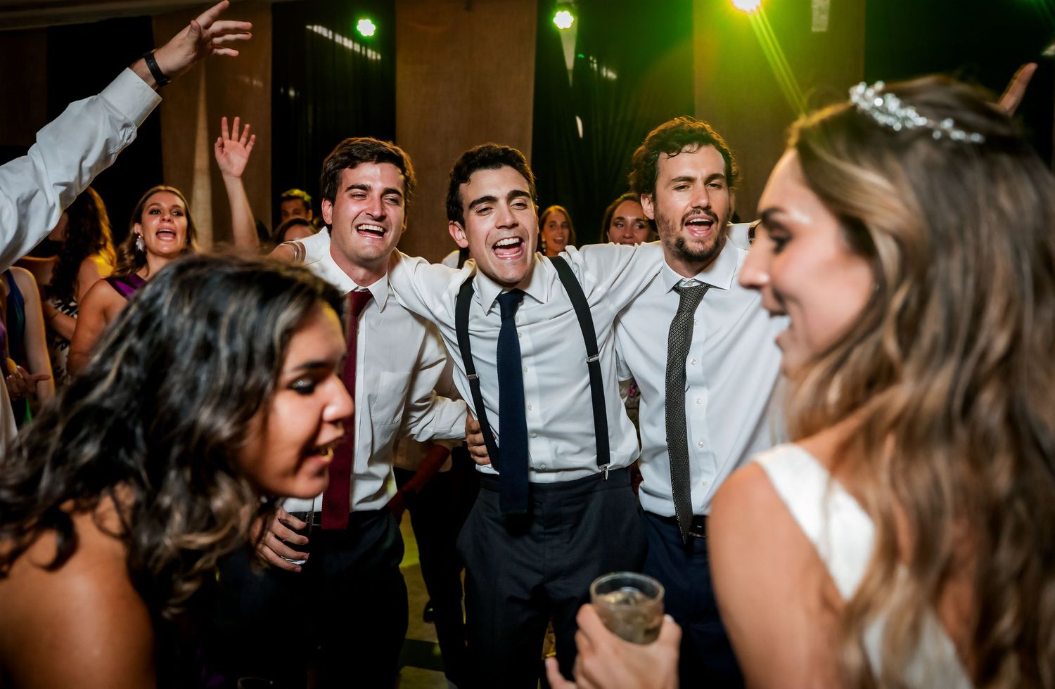 Excited groomsmen in suspenders celebrating with the bride at a lively wedding reception party.