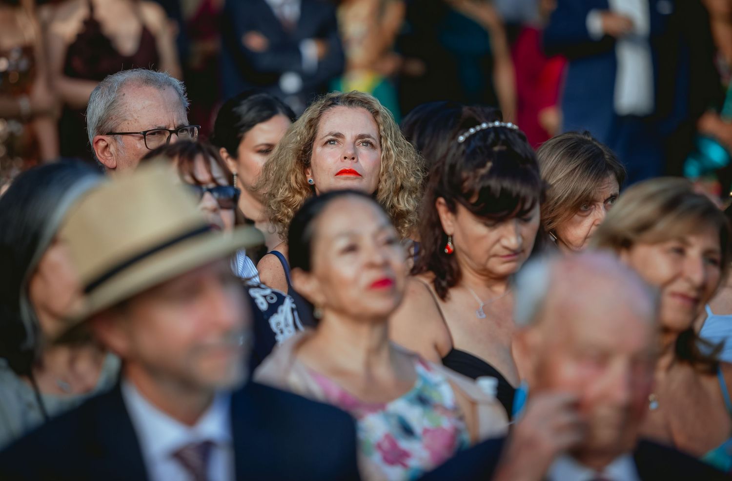 Crowd of elegantly dressed wedding guests watching outdoor ceremony, woman with curly hair visible in center.