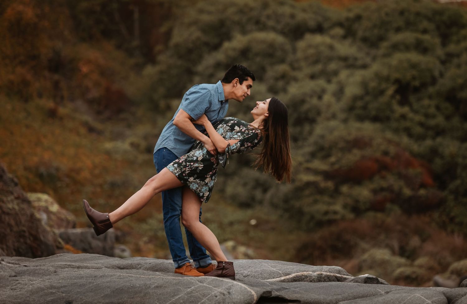 Couple sharing a romantic dip pose on coastal rocks with lush green hillside behind them.