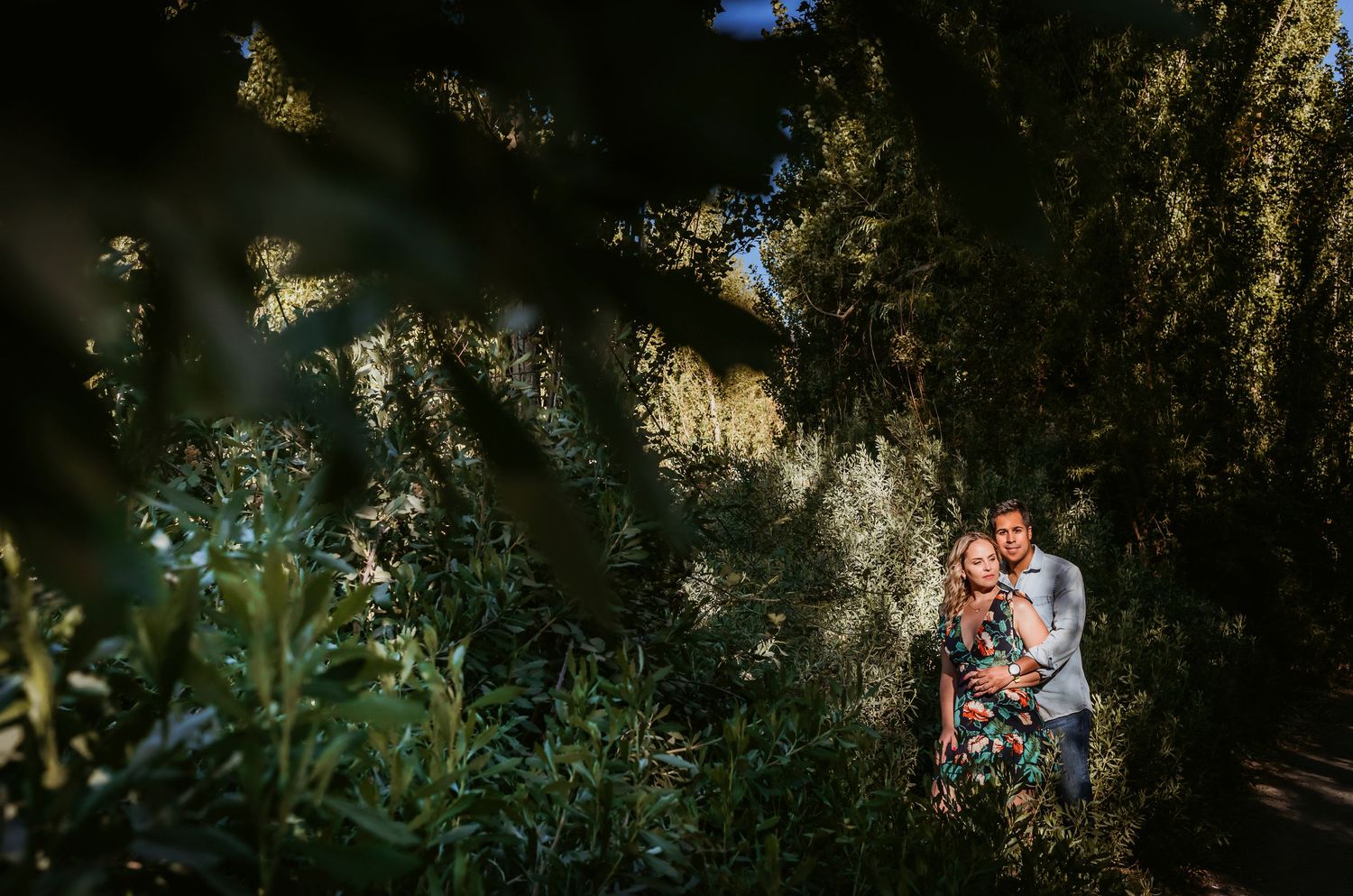Couple embracing romantically in a lush green garden surrounded by tall trees and dappled sunlight.