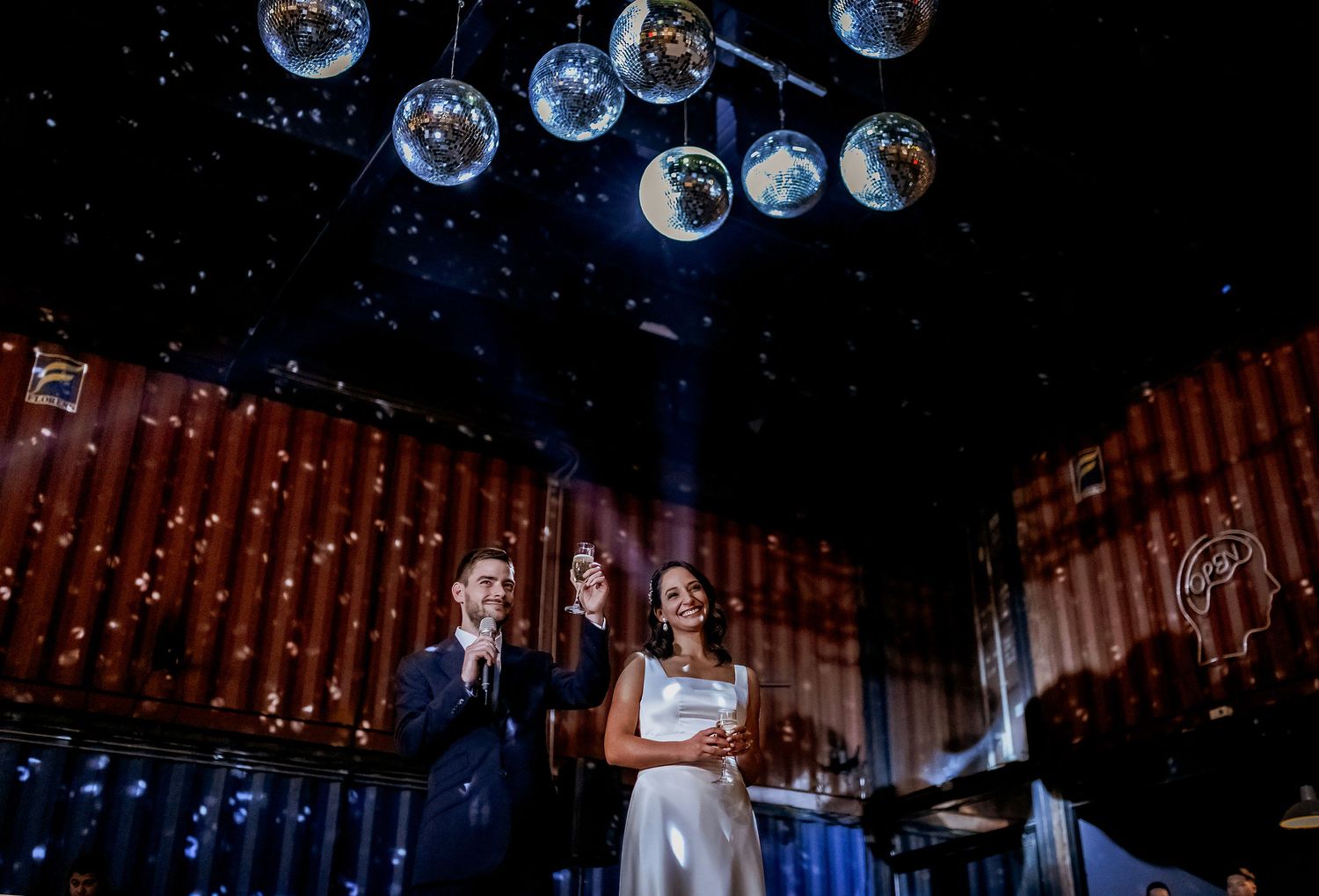 Couple celebrates under disco balls at a festive wedding reception with sparkling lights and red container walls.