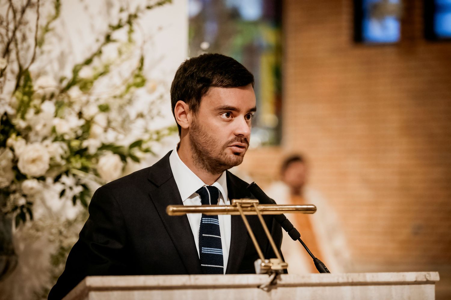 Young man in suit speaking at a podium during a wedding ceremony with white floral arrangements.