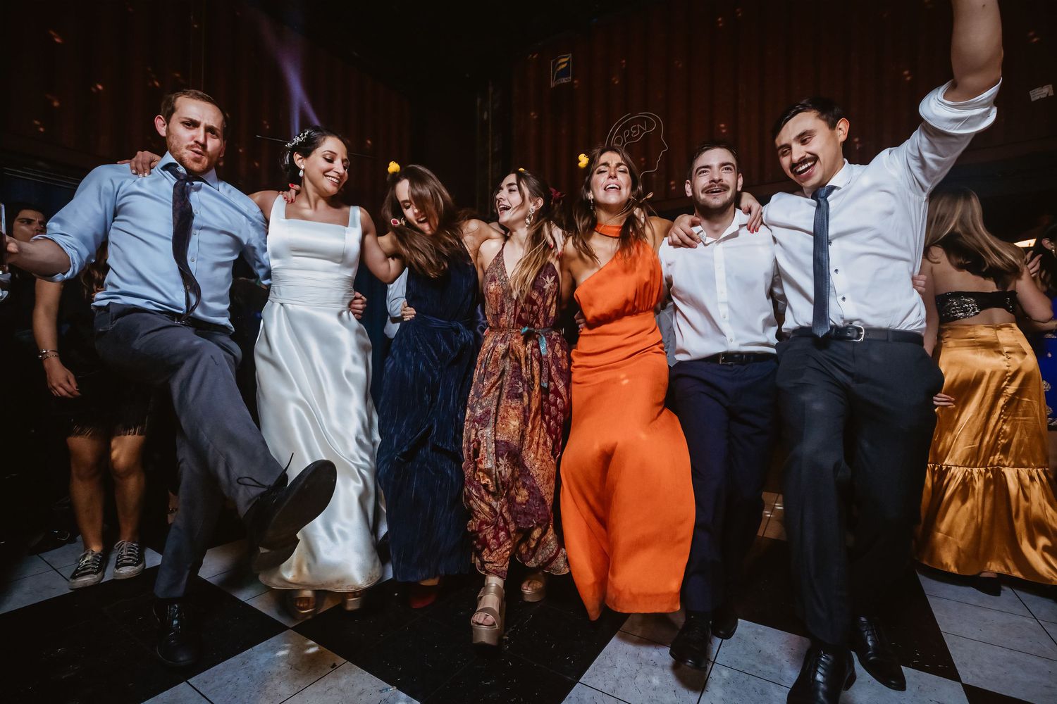 Six wedding guests jumping joyfully on dance floor, bride in white gown surrounded by friends in formal attire.