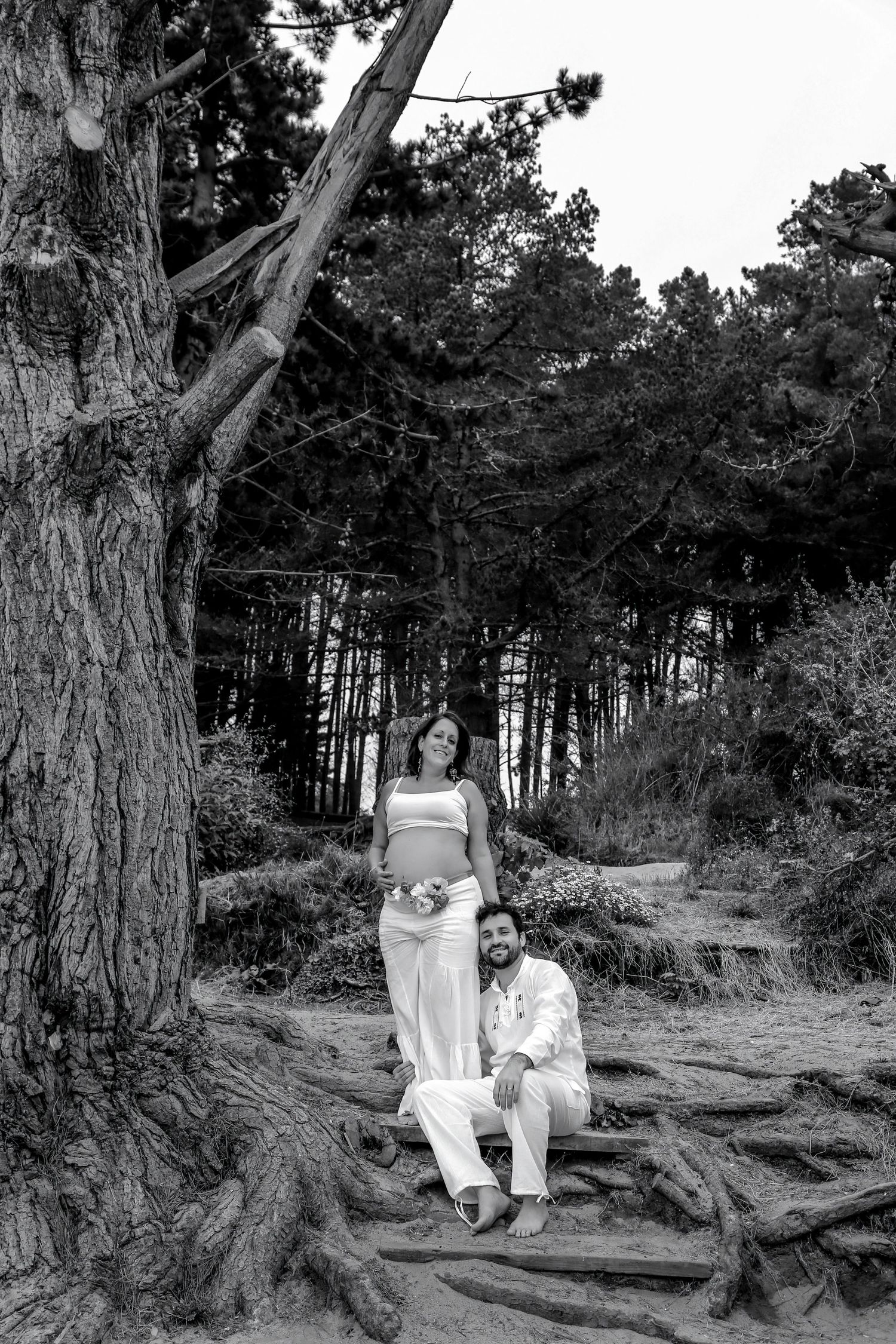 Black and white photo of a pregnant couple posing outdoors among tall trees on wooden steps.