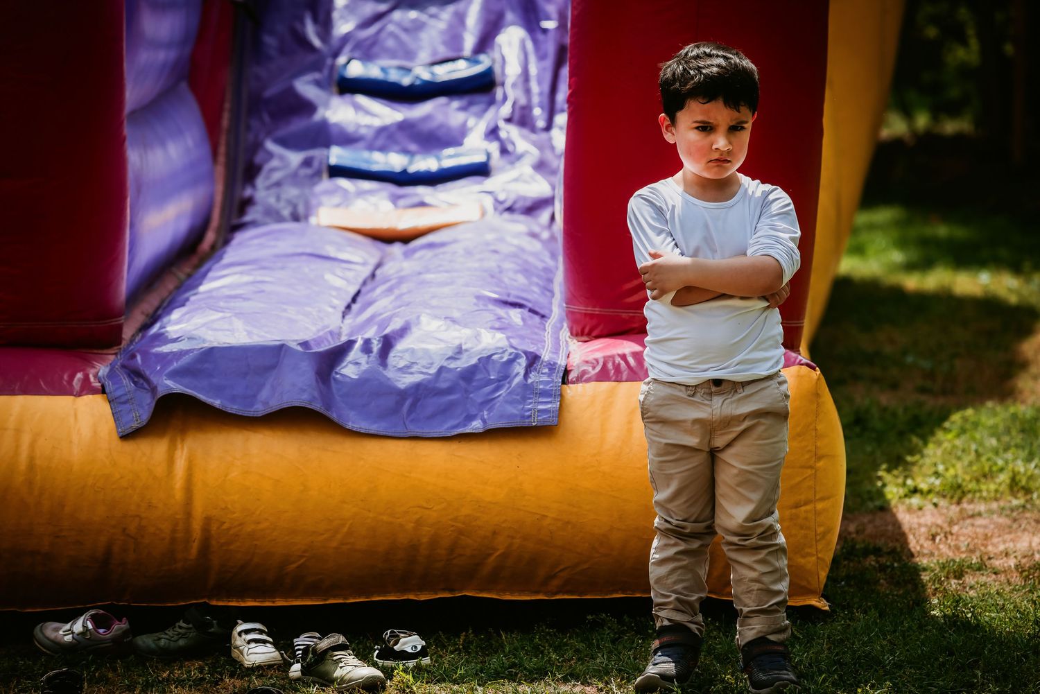 Young boy with crossed arms standing beside a colorful inflatable bounce house at an outdoor party.