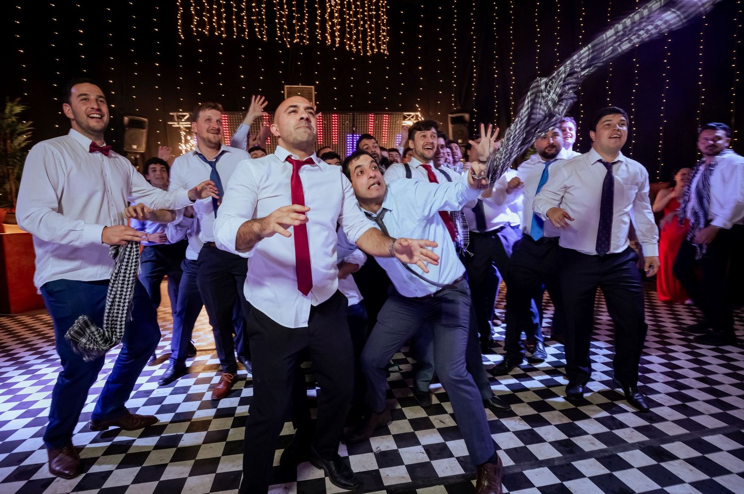 Groomsmen in white shirts and ties reach for bouquet toss on checkered wedding dance floor.