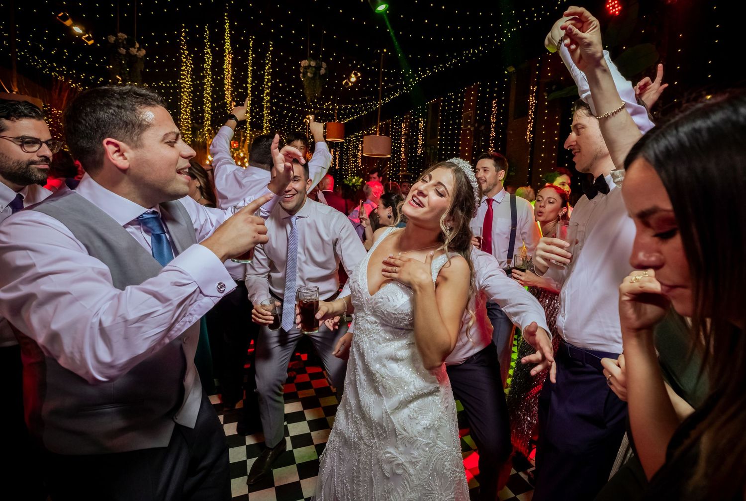 Bride and groom dancing joyfully with guests at a lively wedding reception on a checkered dance floor.