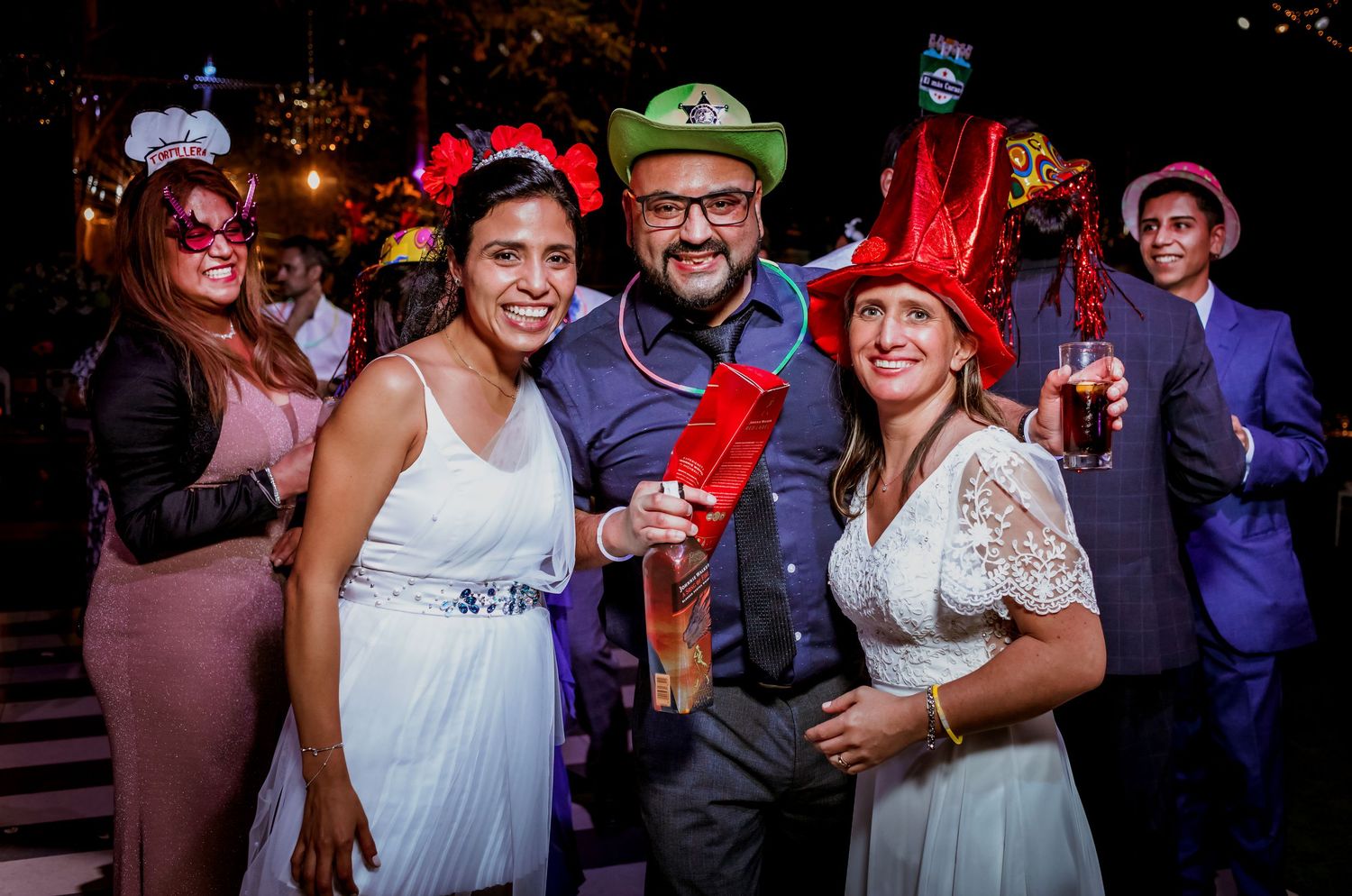 Two brides in white dresses pose with a male guest, all wearing colorful party hats at wedding reception.