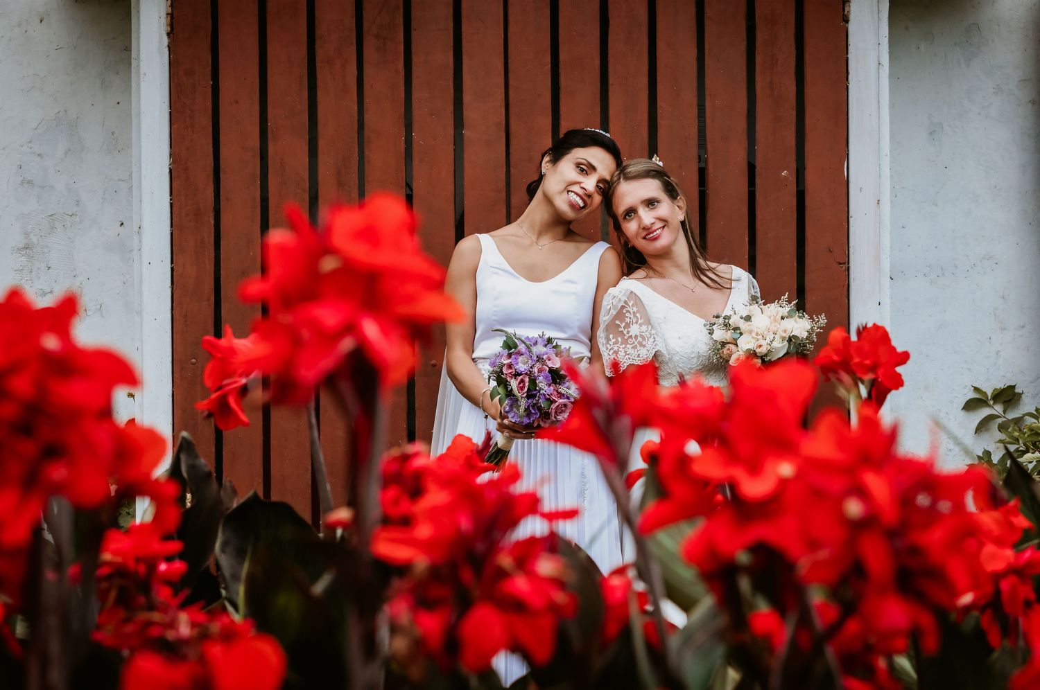 Two brides leaning together affectionately holding bouquets in front of rustic red wooden door.