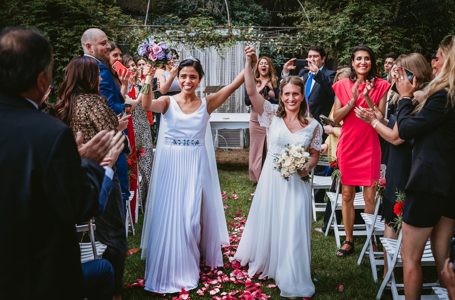 Two brides walk triumphantly down the aisle with hands raised after their wedding ceremony.