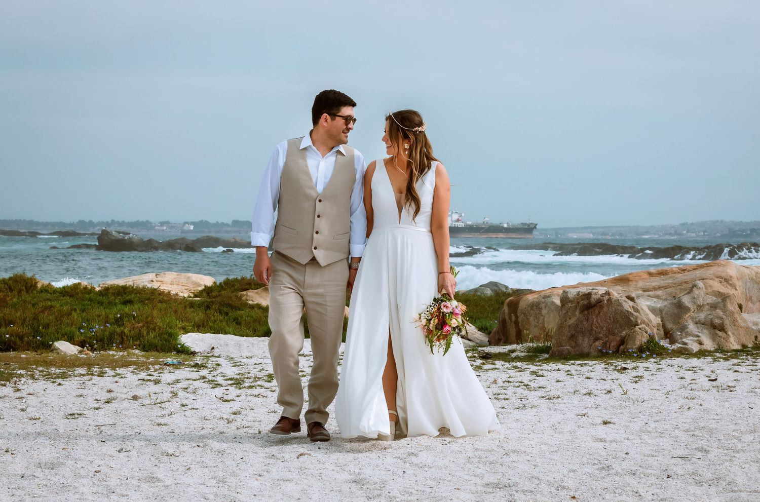 Bride and groom walking hand in hand on sandy beach with ocean waves and rocks in the background.
