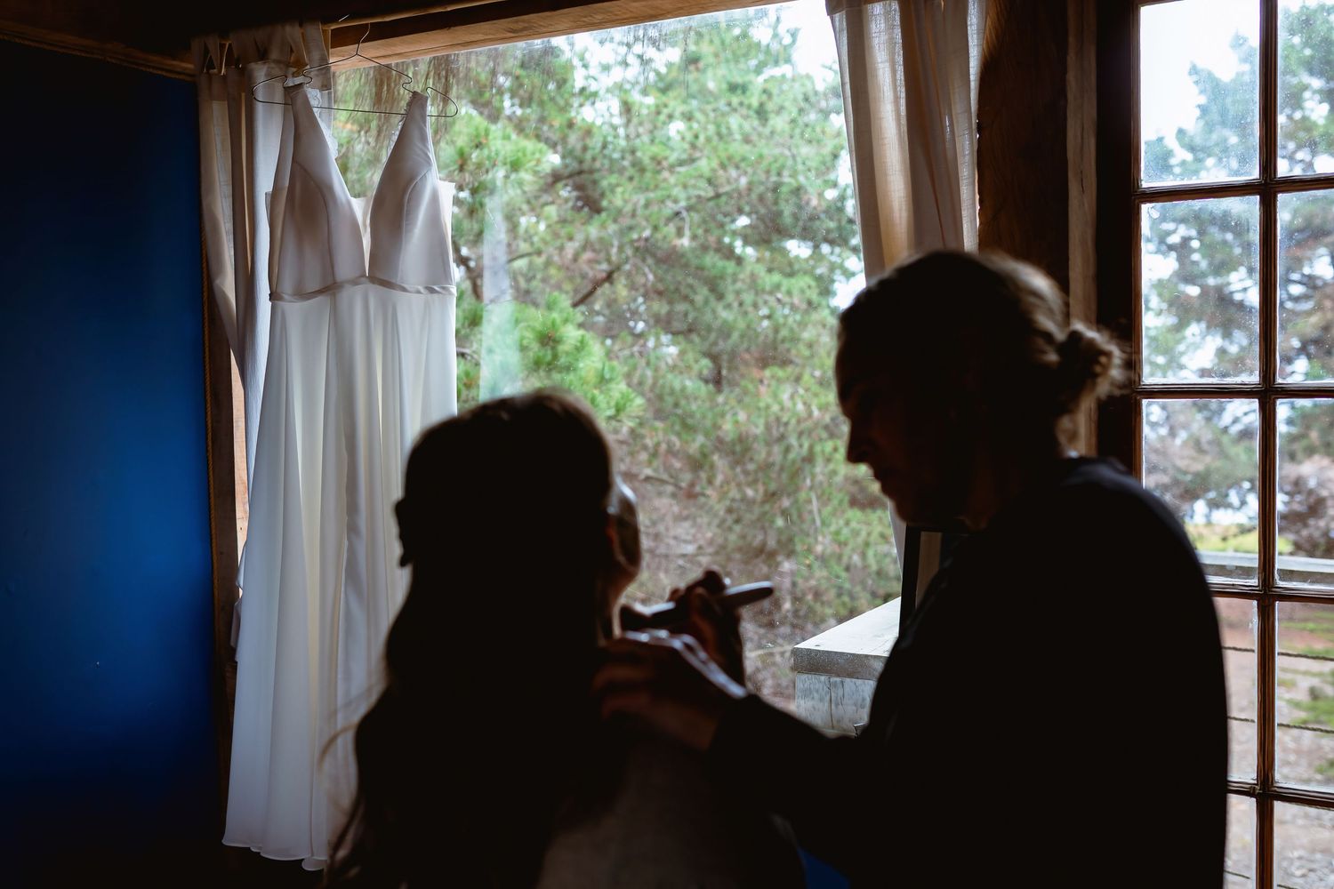 Bride getting makeup done in silhouette near window with wedding dress hanging in background.