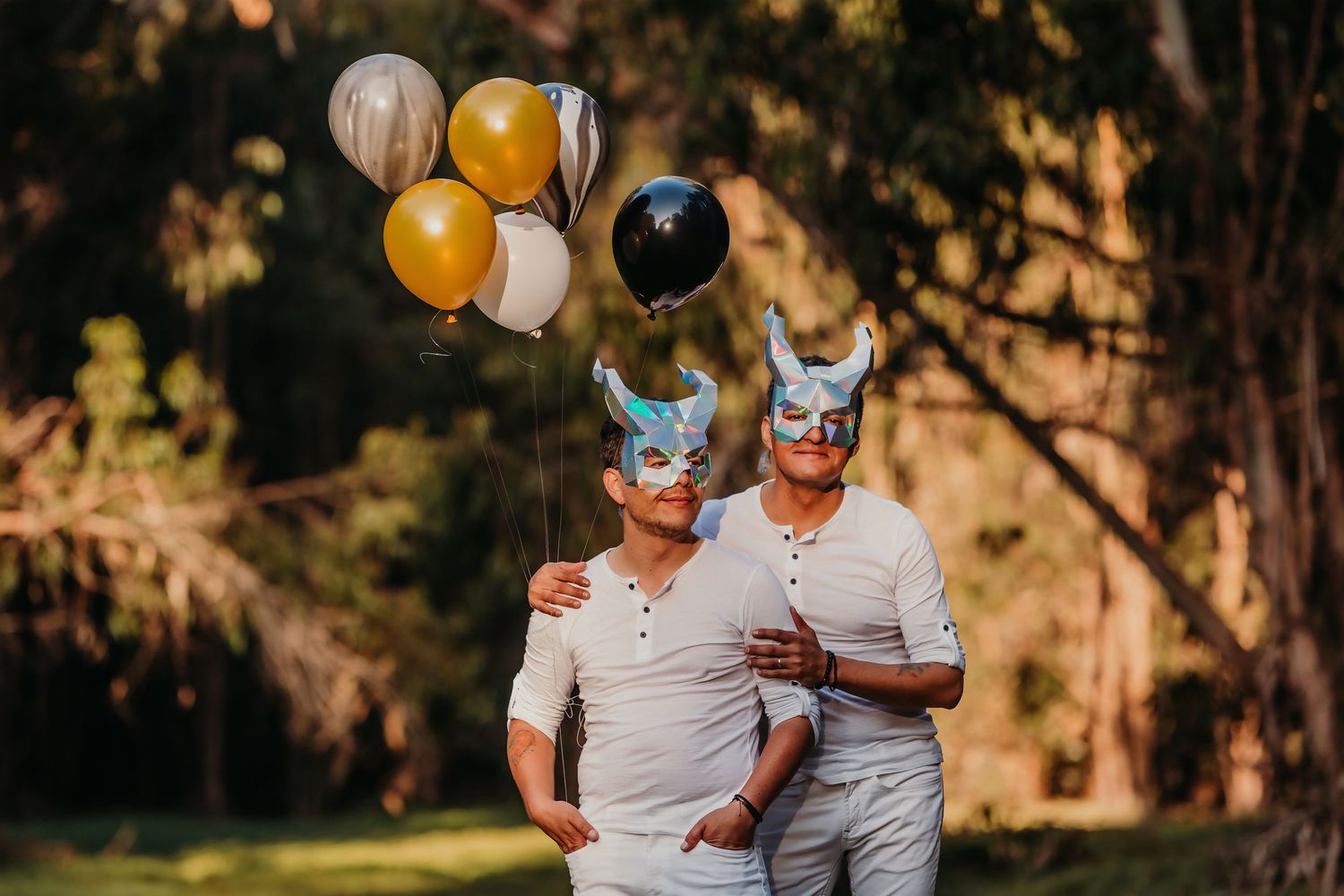 Two men in white outfits and Viking masks pose together holding gold and silver balloons in a park.