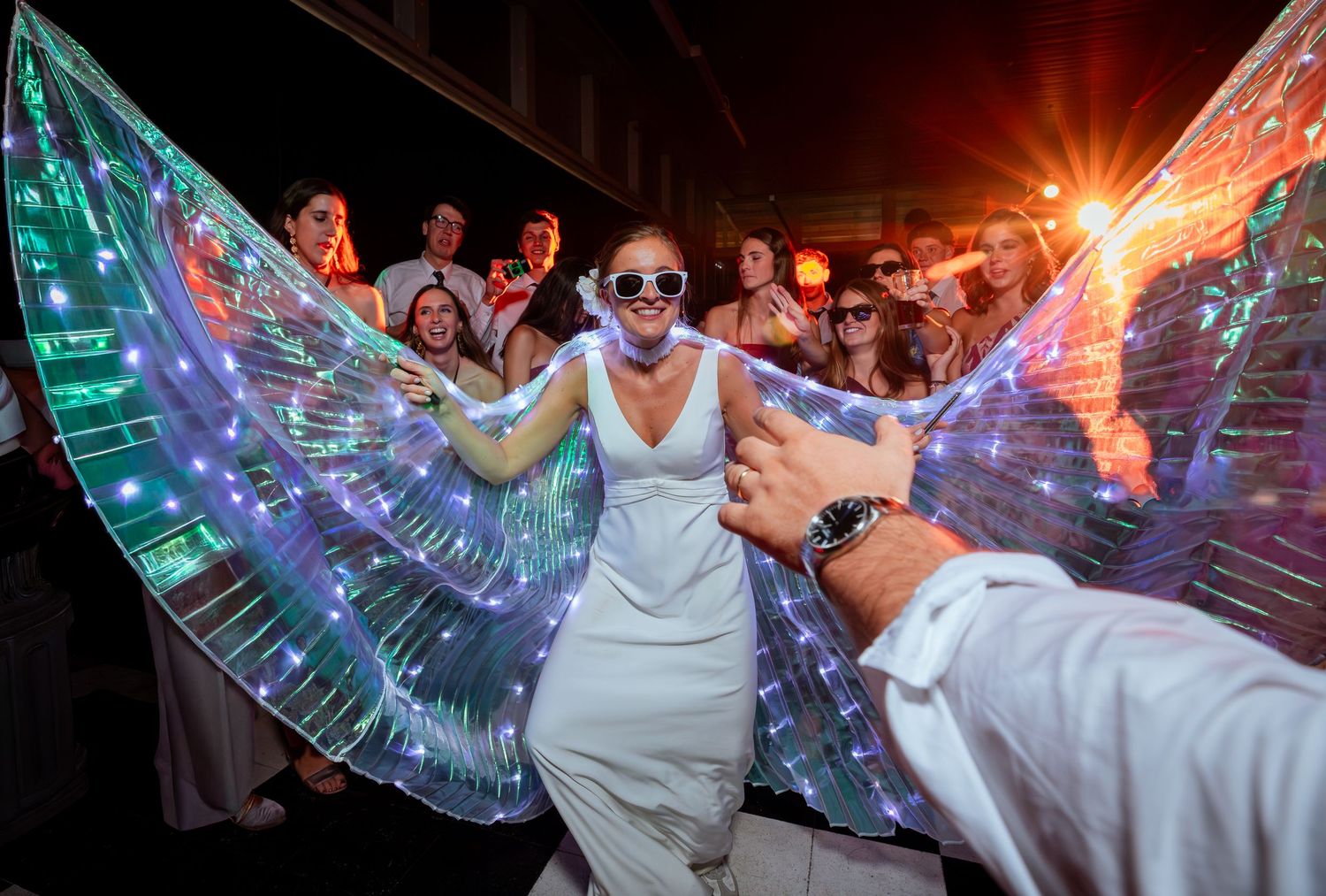 Bride wearing glowing LED butterfly wings surrounded by cheering guests at wedding reception.