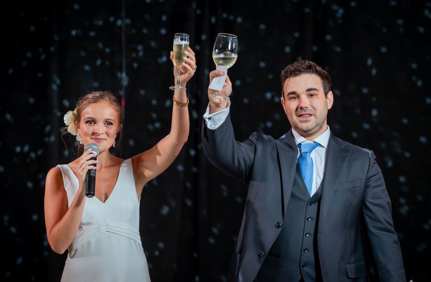 Bride and groom raising champagne glasses in toast while bride speaks into microphone at wedding.