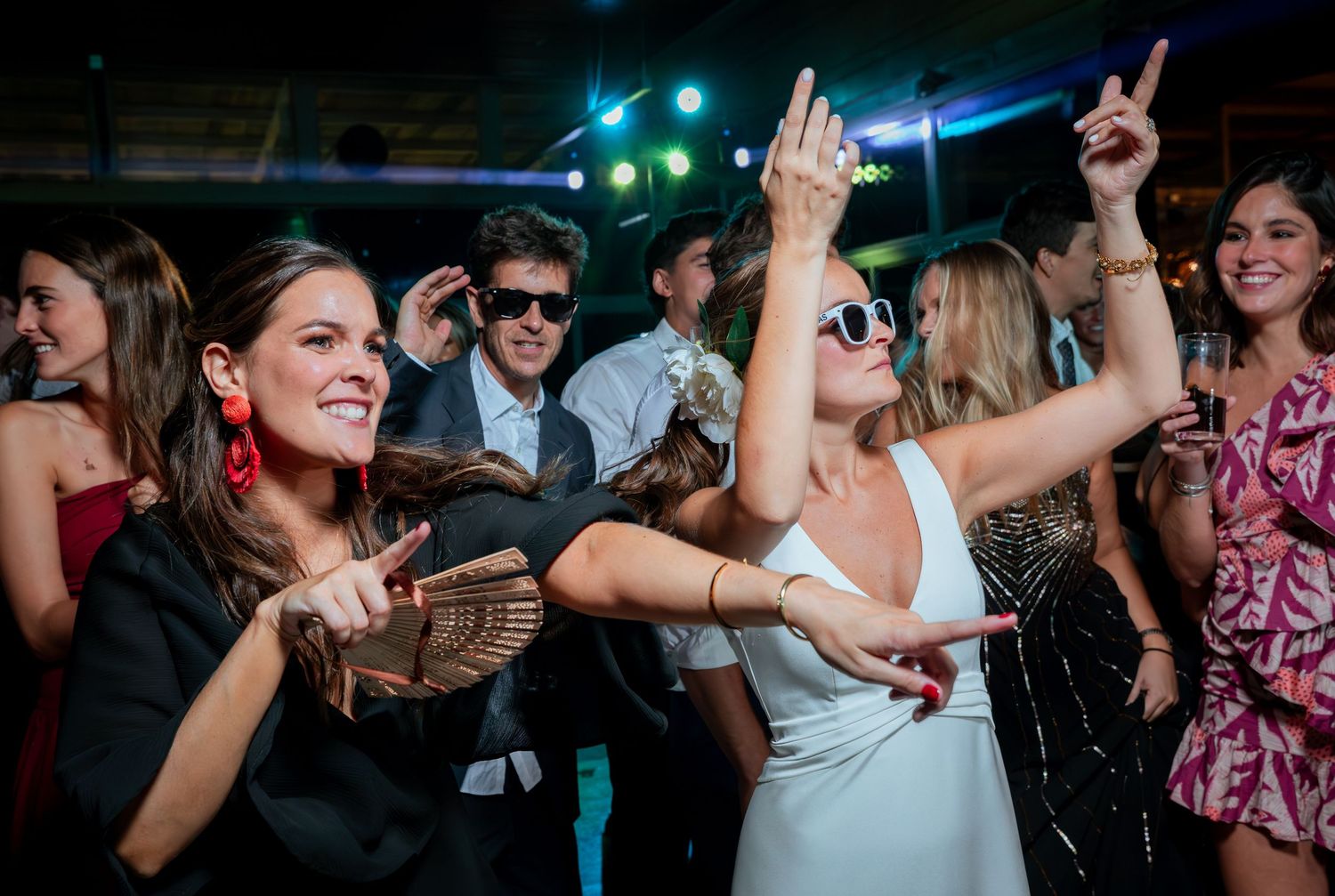 Bride in white dress wearing sunglasses dances with friend at vibrant wedding reception.
