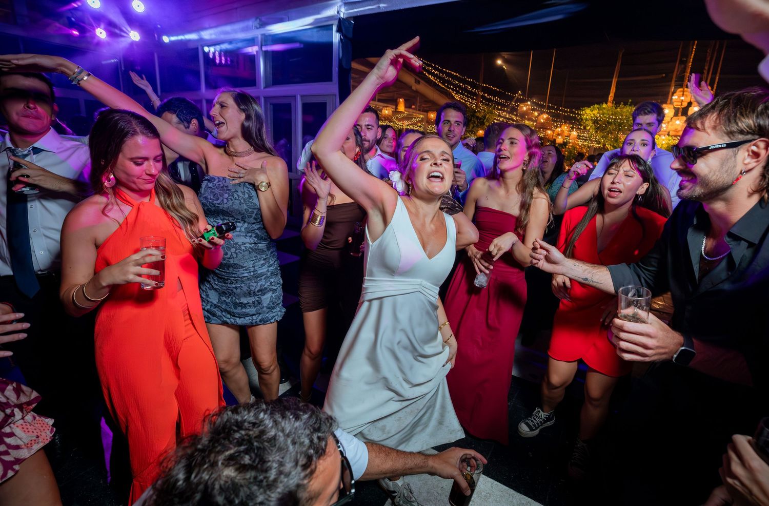 Bride in white halter dress dances with arms raised among colorfully dressed guests at outdoor wedding reception.