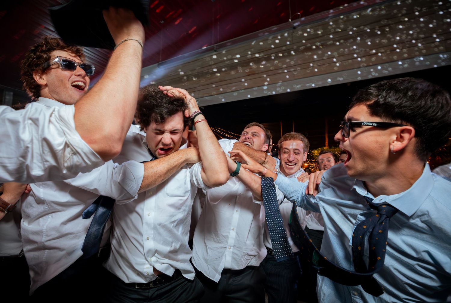 Groomsmen in white shirts and ties laugh and roughhouse at wedding reception.