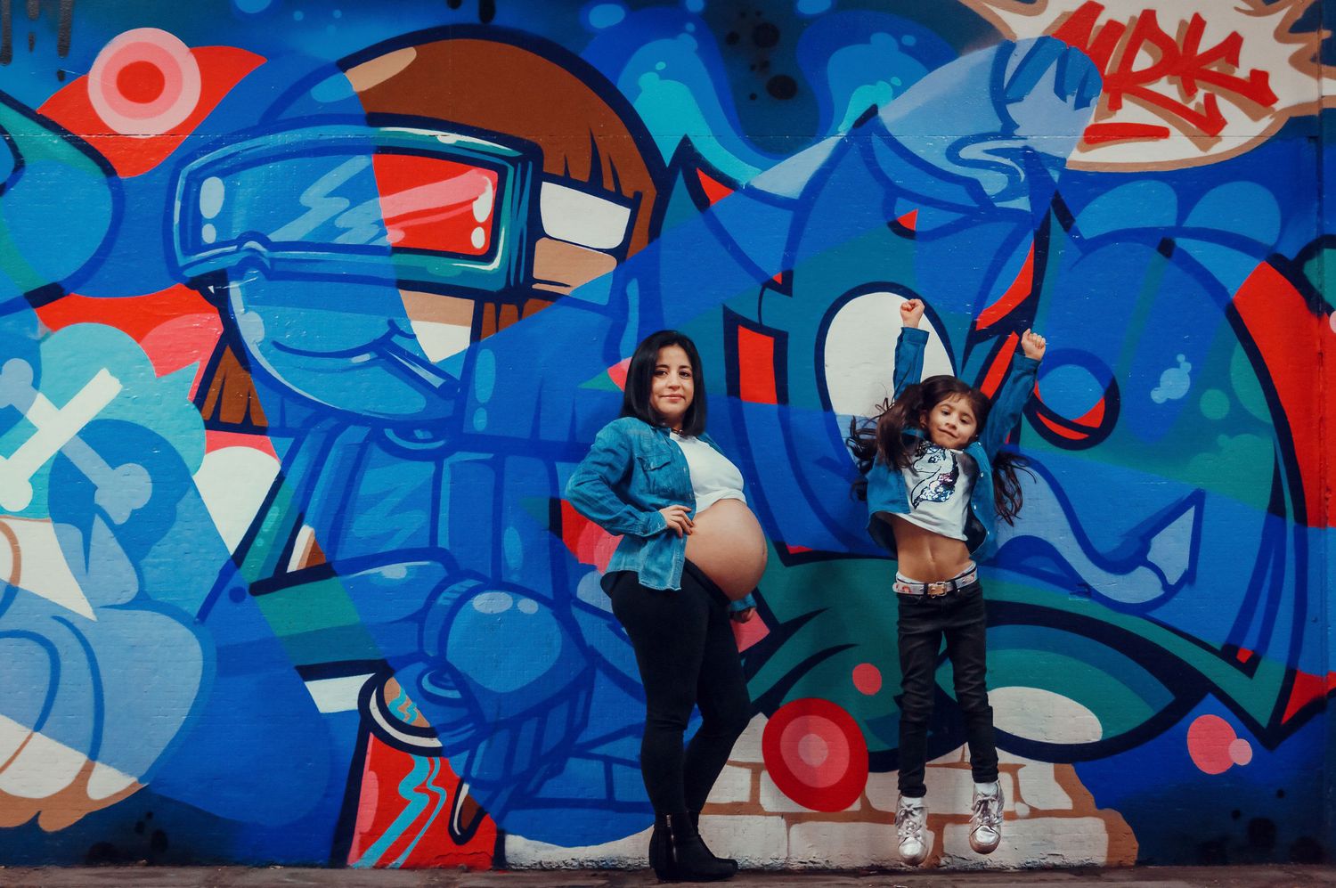 Pregnant woman and young girl posing energetically in front of a colorful blue graffiti wall.