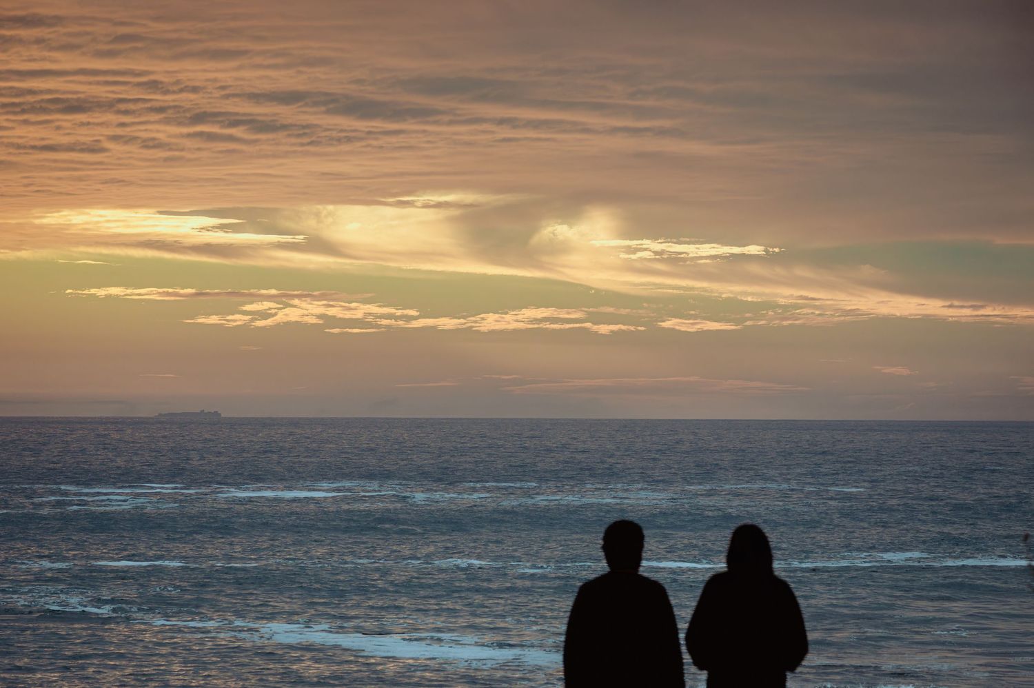 Two silhouetted figures stand together watching a dramatic ocean sunset with colorful clouds.