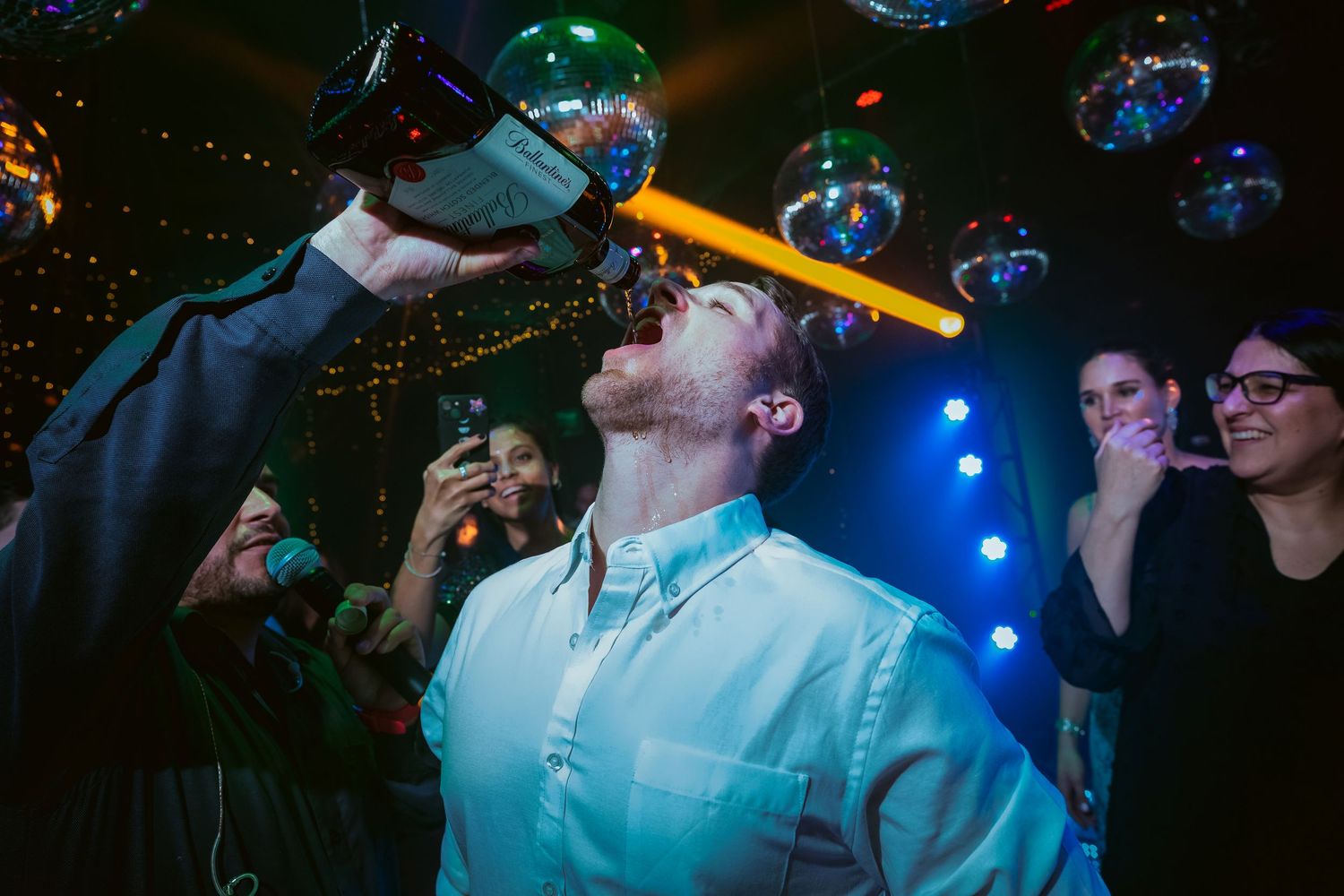 Man drinking from a bottle at a lively party with disco balls and colorful lights in the background.