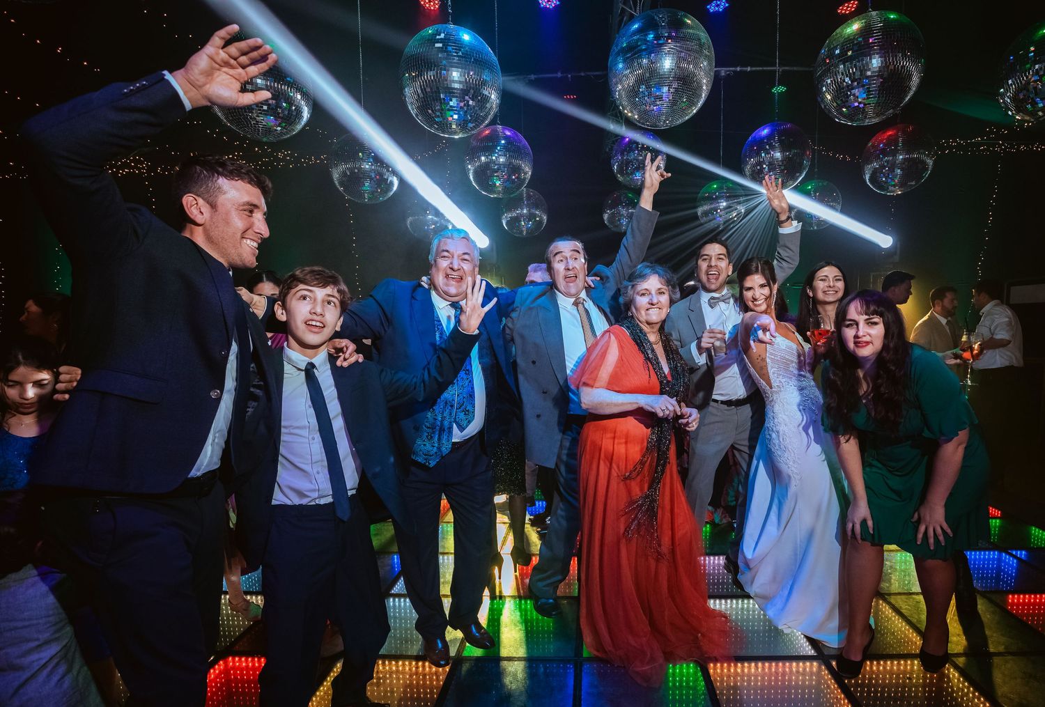 Group of elegantly dressed people dancing under disco balls on a colorful LED dance floor at a festive party.