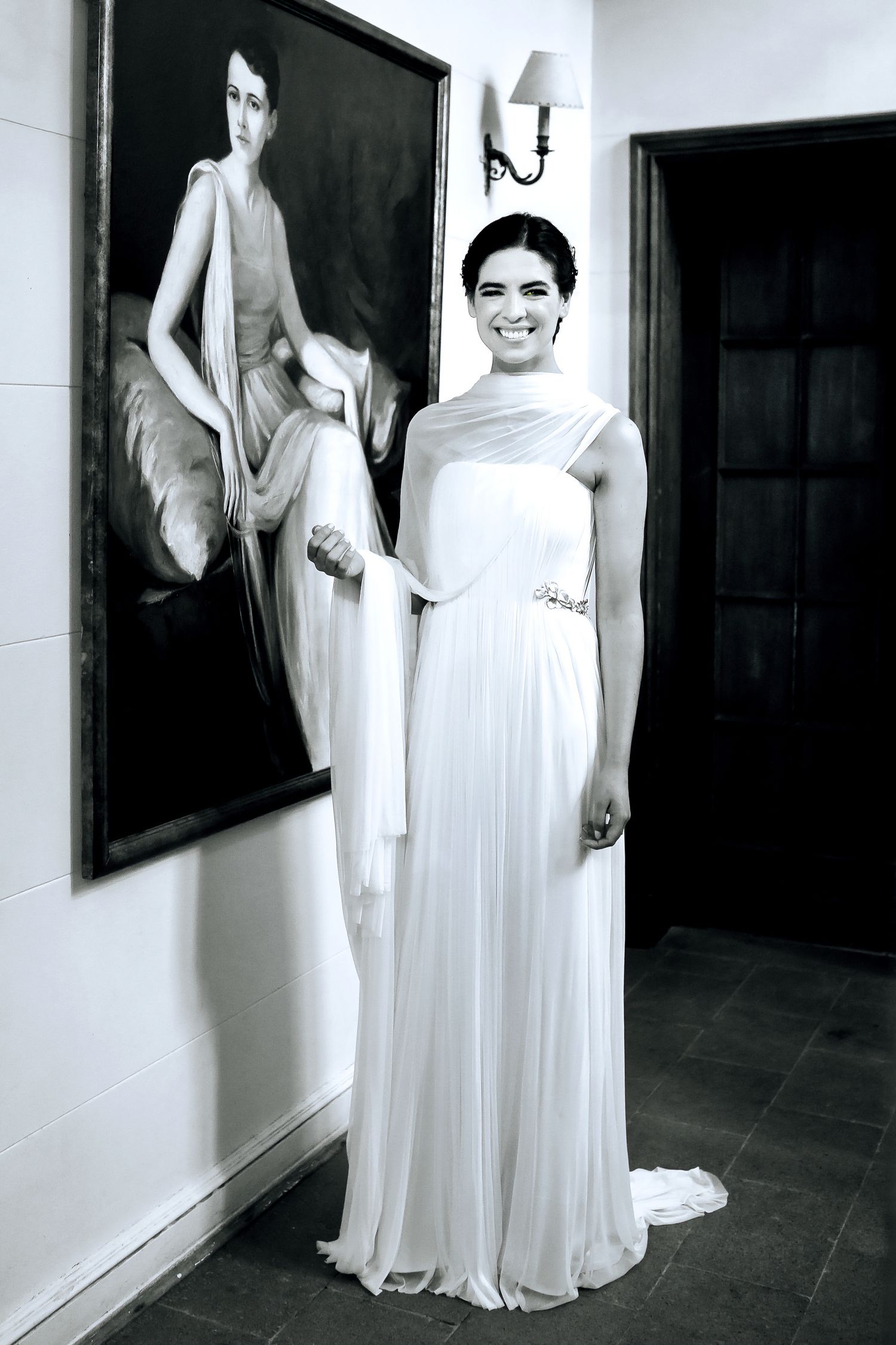 Elegant bride in white Grecian gown smiling beside classic portrait painting in black and white photo.