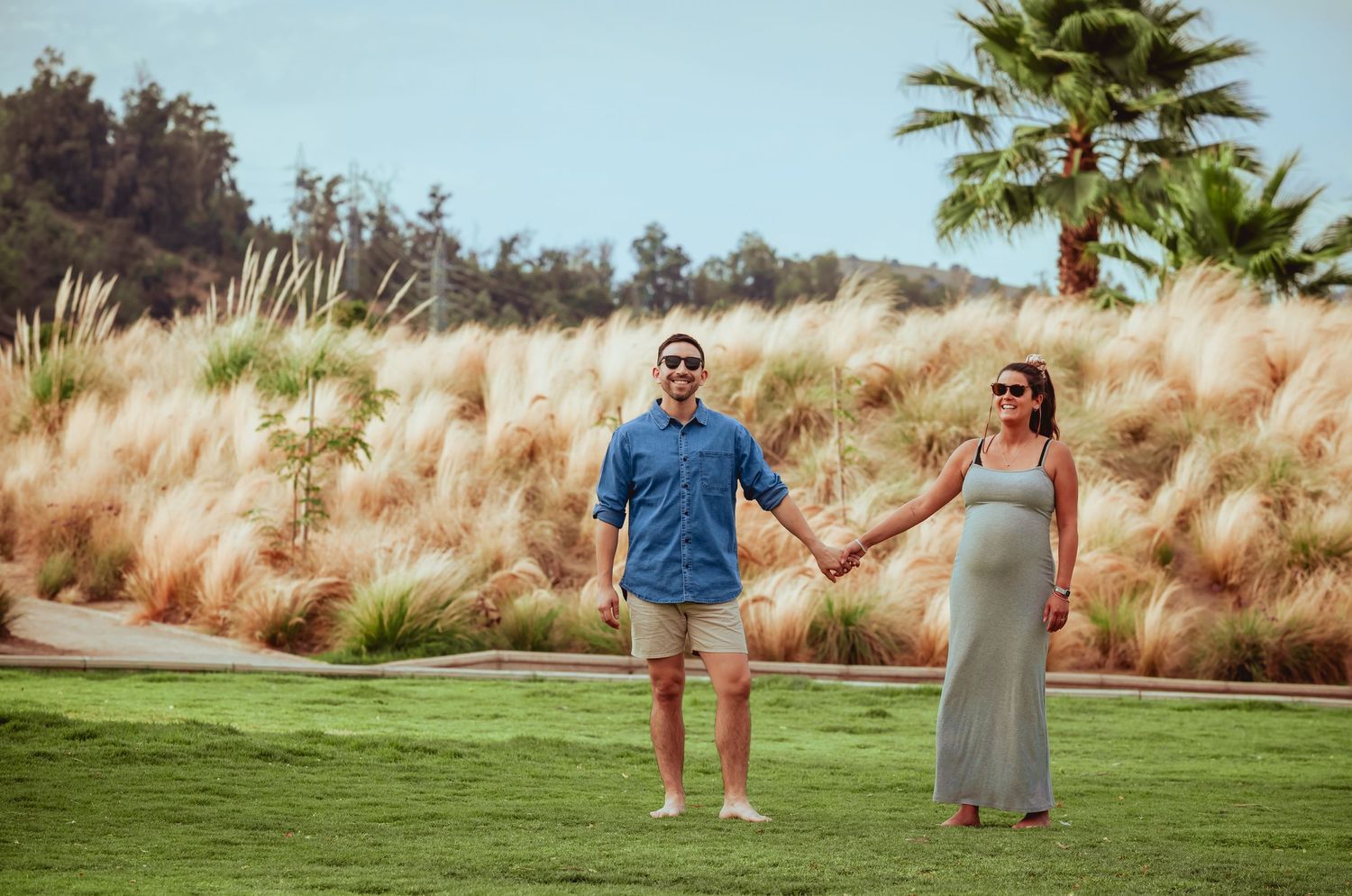 Pregnant couple holding hands on lush green lawn with tall golden pampas grass and palm trees.