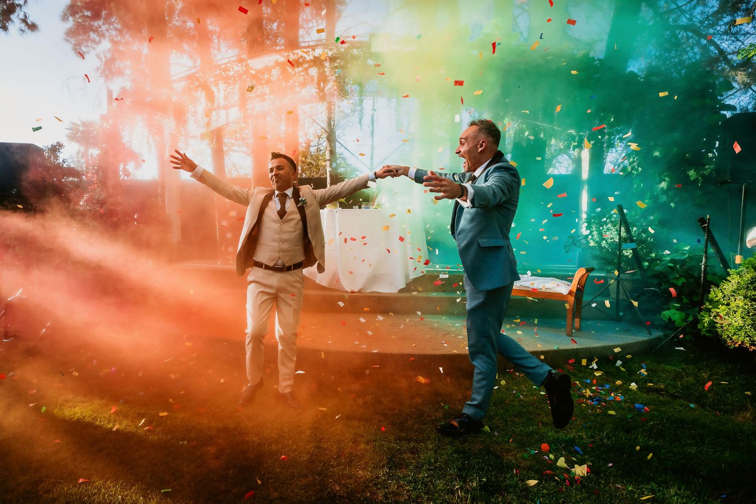 Two men celebrating outdoors with vibrant rainbow colored smoke bombs and confetti in a garden setting.