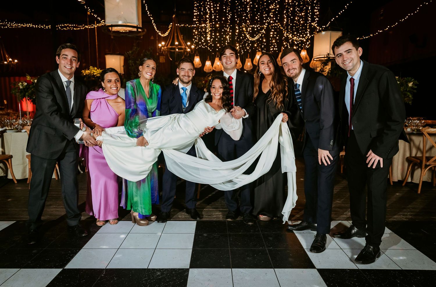 Group of elegantly dressed guests posing on checkered dance floor at a wedding reception with fairy lights.