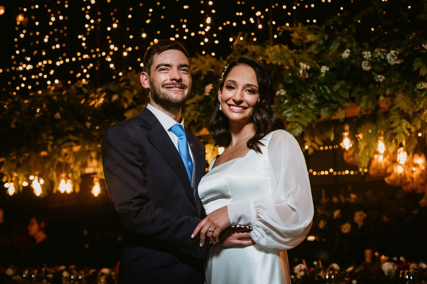 Smiling couple in elegant wedding attire posing together under warm fairy lights and lush greenery backdrop.