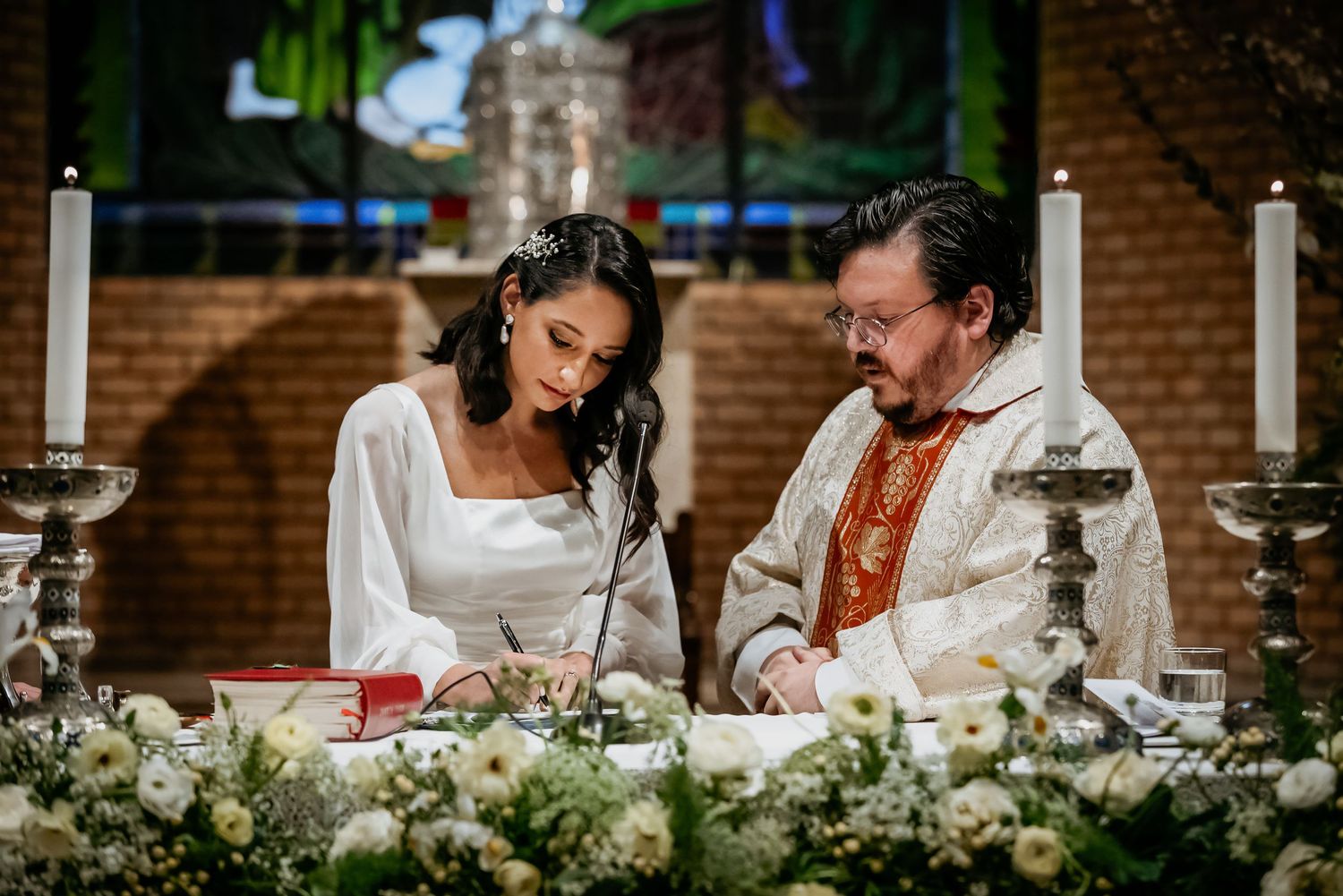 Bride in white gown and priest in red vestments signing wedding documents at flower-adorned altar in church.