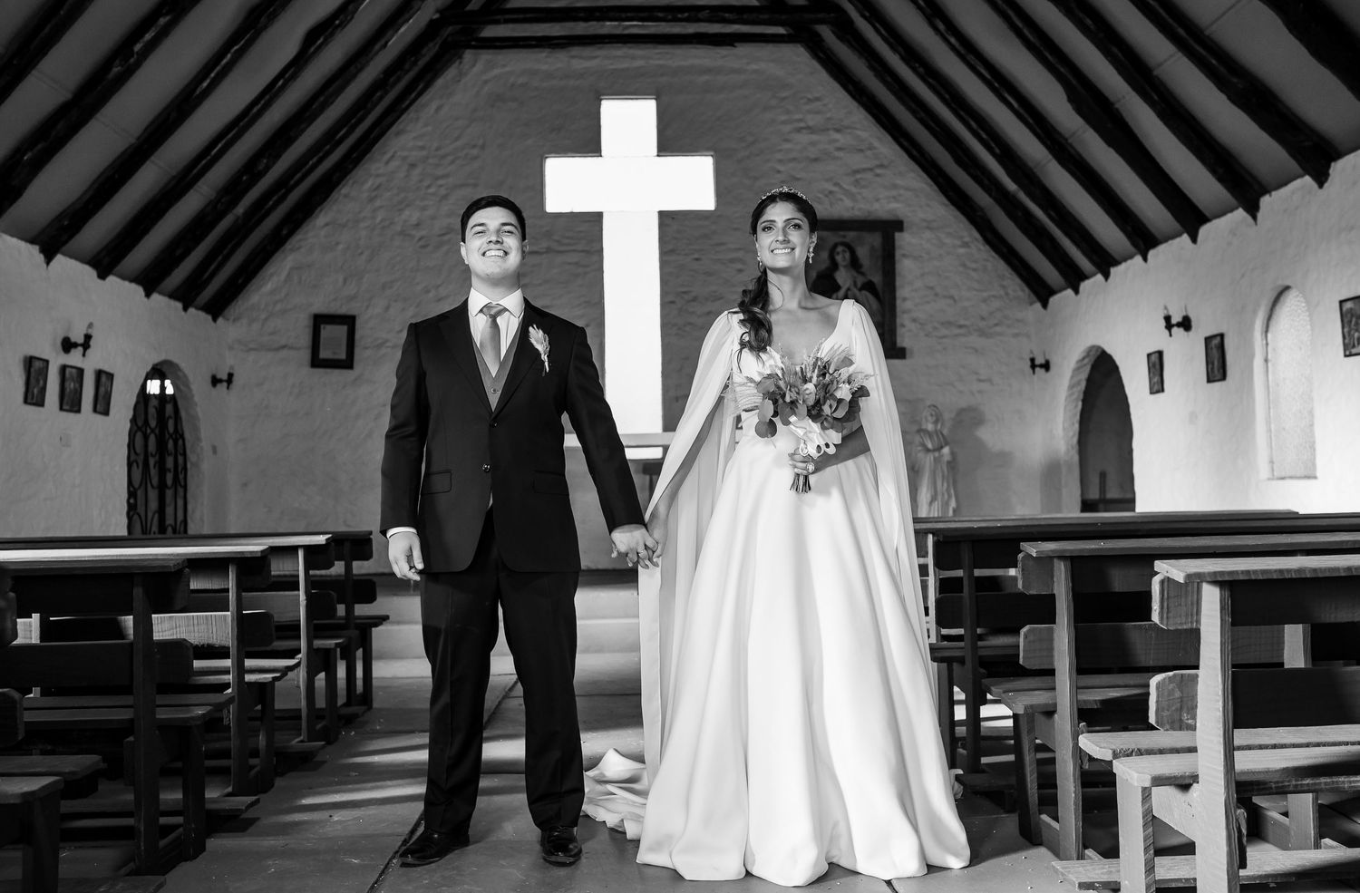 Black and white photo of a bride and groom holding hands in a church aisle with a large cross behind them.