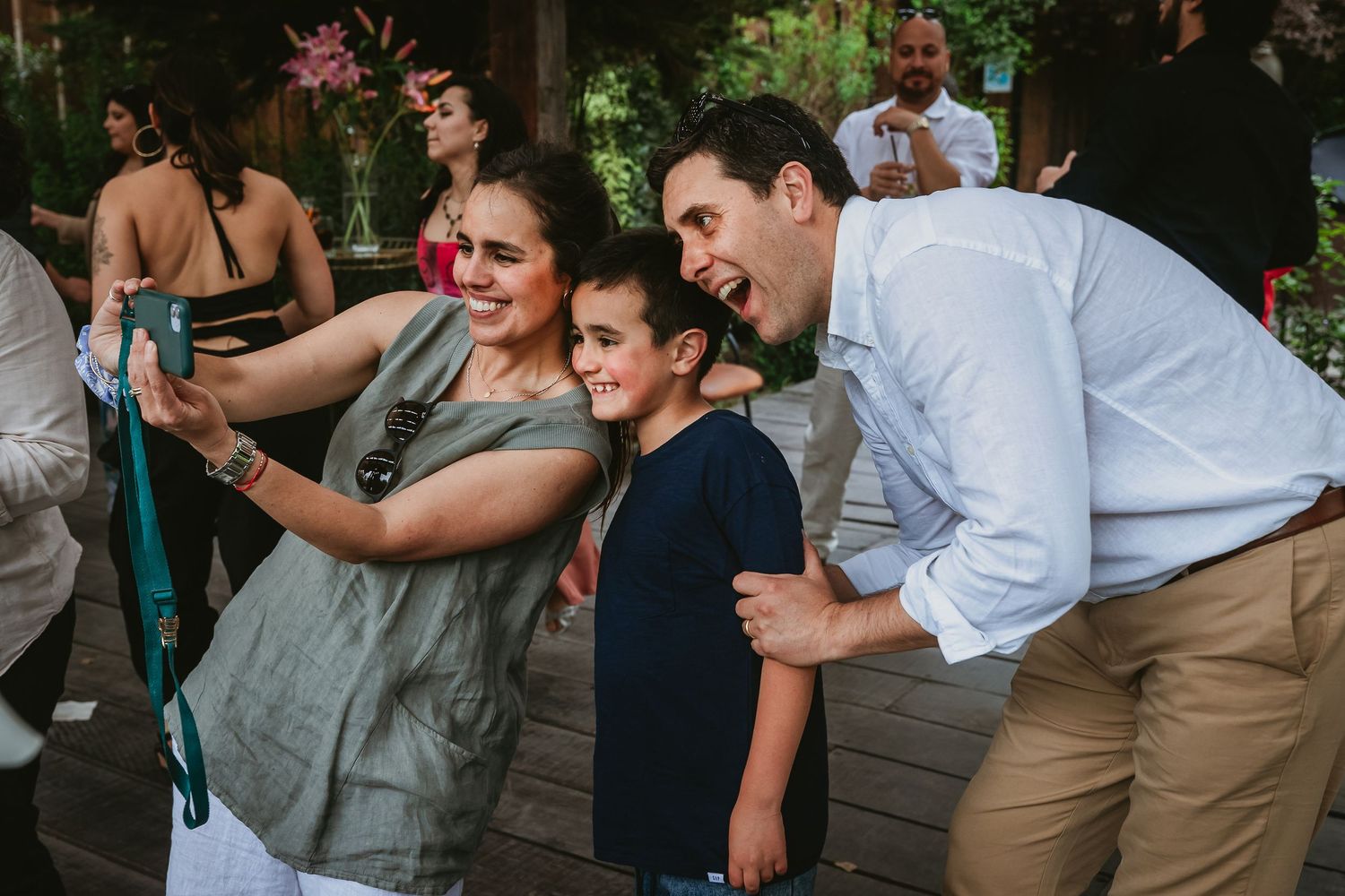 Woman and boy take selfie with smiling man at outdoor wedding reception celebration.