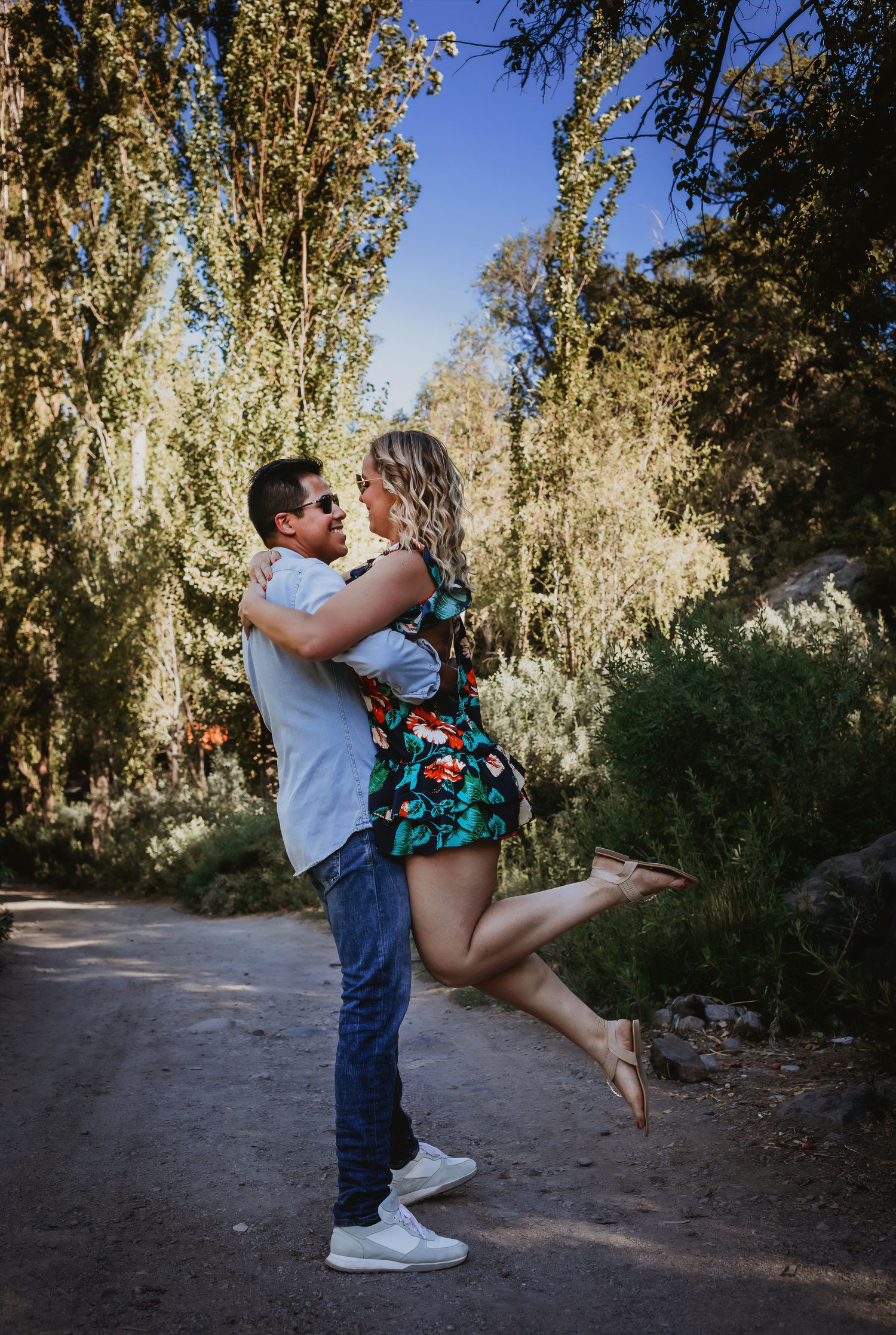 A man lifts a woman in a floral dress on a sunny tree-lined path, sharing a romantic moment outdoors.