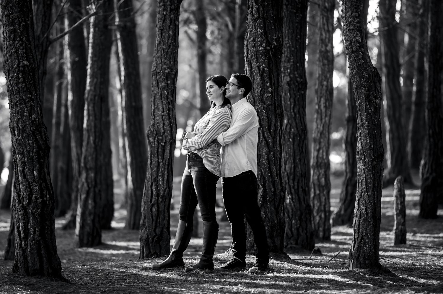 Black and white photo of a couple standing among tall trees in a forest, the man embracing the woman from behind.