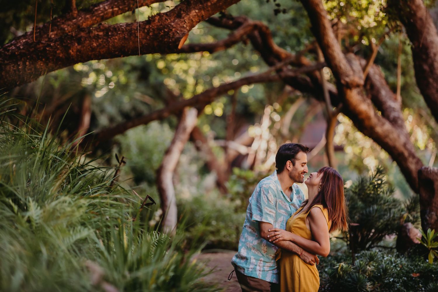 Couple sharing a romantic kiss surrounded by lush tropical trees and greenery in a botanical garden.