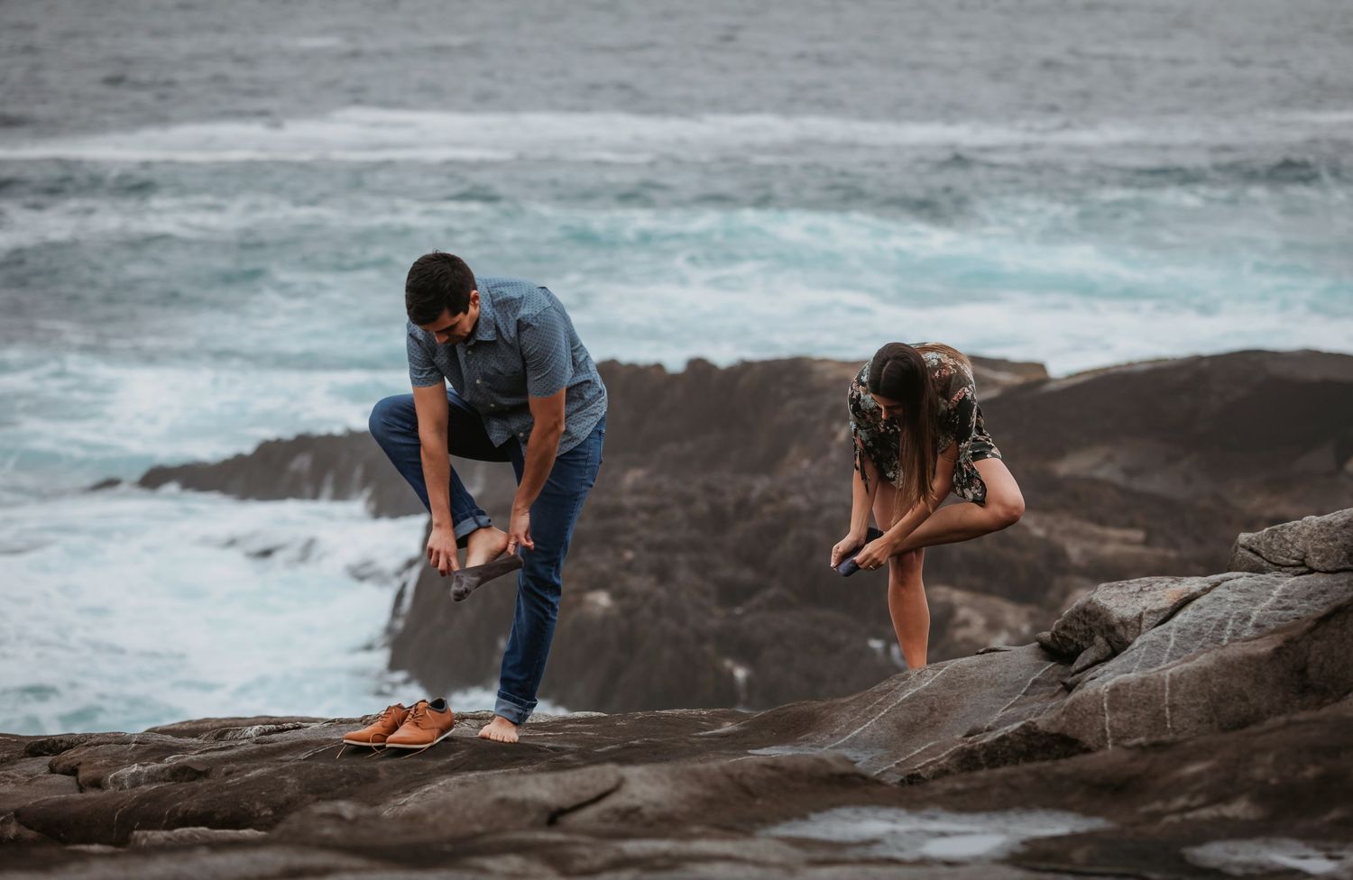 Couple helping each other navigate rocky coastal terrain with blue ocean waves in the background.